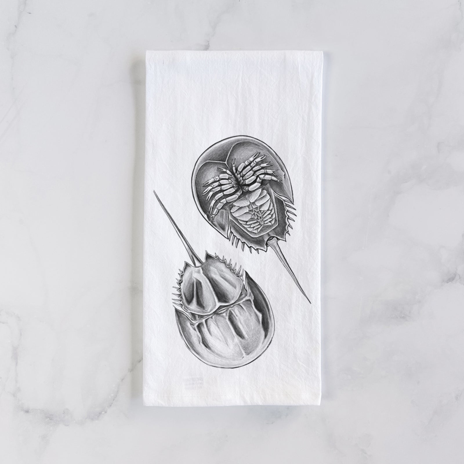 White cotton tea towel printed with a detailed black ink illustration of two horseshoe crabs, on a marble counter beside a round wooden cutting board and greenery.