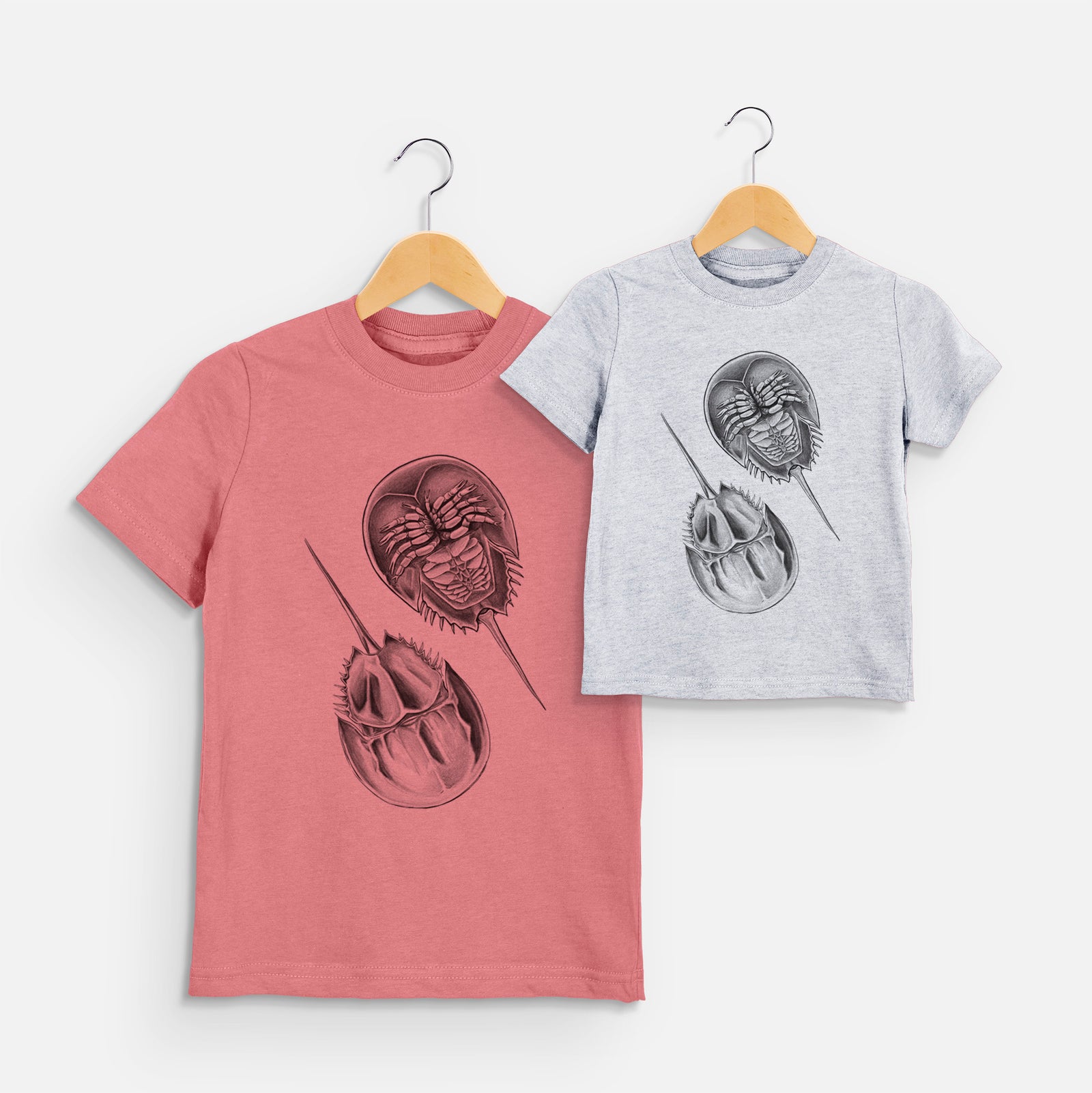 Two youth t-shirts in different colors on hangers, printed with a detailed black ink illustration of two horseshoe crabs.