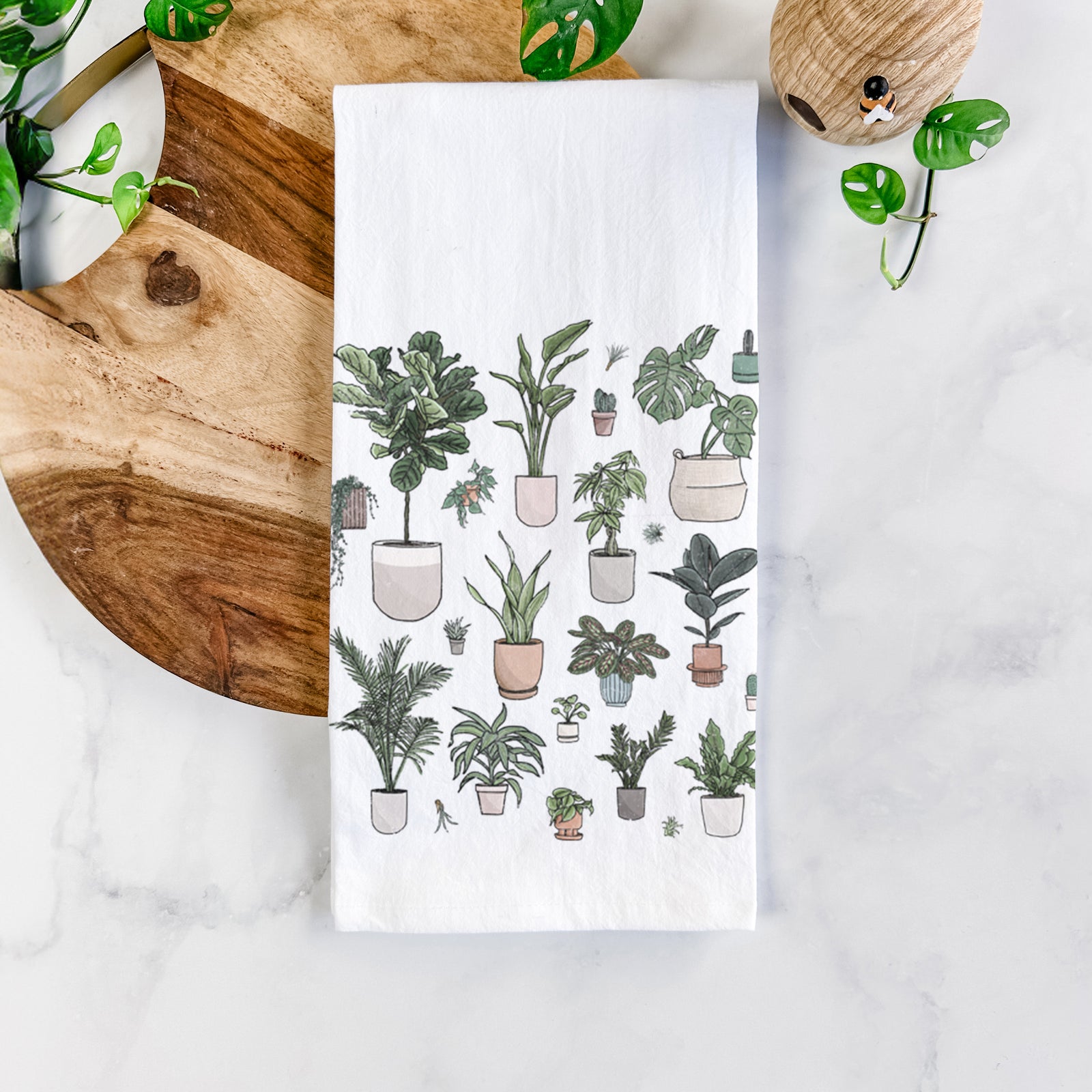White cotton tea towel printed with a full-color collection of potted houseplants in different pots, on a marble counter beside a round wooden cutting board and greenery.