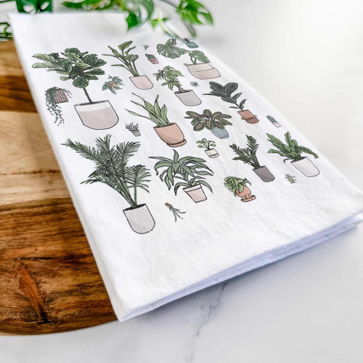 Close-up of a folded white tea towel on a wooden board, showing a full-color collection of potted houseplants in different pots.