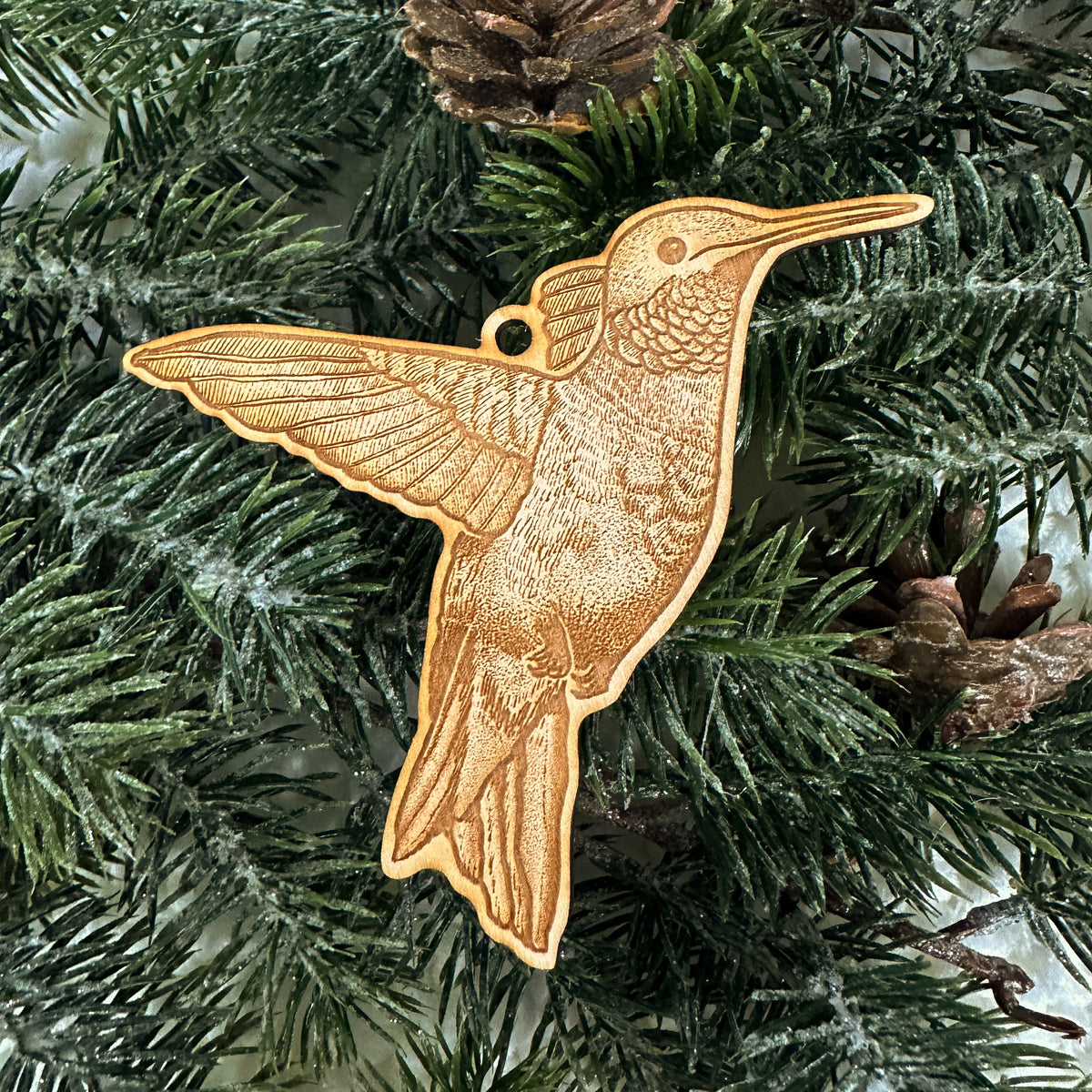 Wooden ornament featuring a detailed engraving of a hummingbird in flight, on a knit blanket with pine cones.