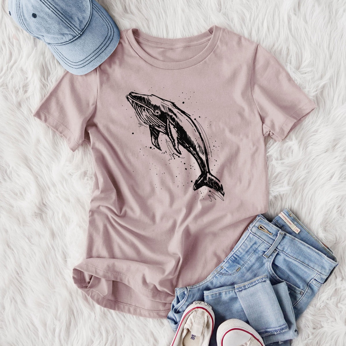 Women's relaxed fit cotton t-shirt printed with a black ink illustration of a humpback whale diving, with ink splatters, styled flat on a white faux fur rug with jeans.
