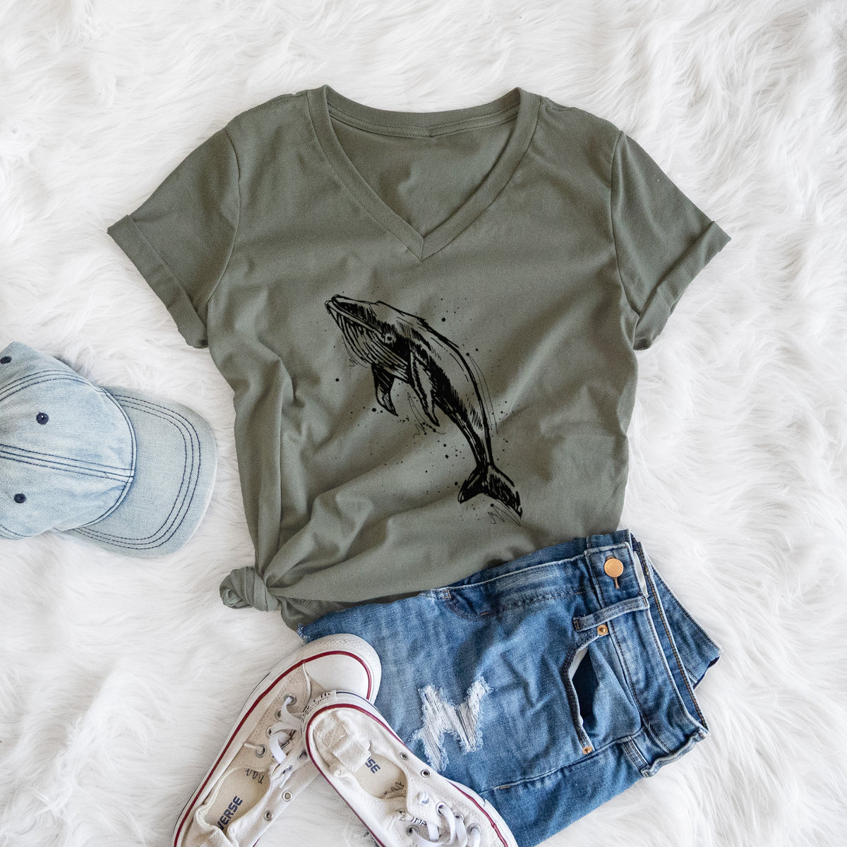 Military green women's relaxed fit cotton V-neck t-shirt printed with a black ink illustration of a humpback whale diving, with ink splatters, styled flat on a white faux fur rug with a cap, jeans and sneakers.