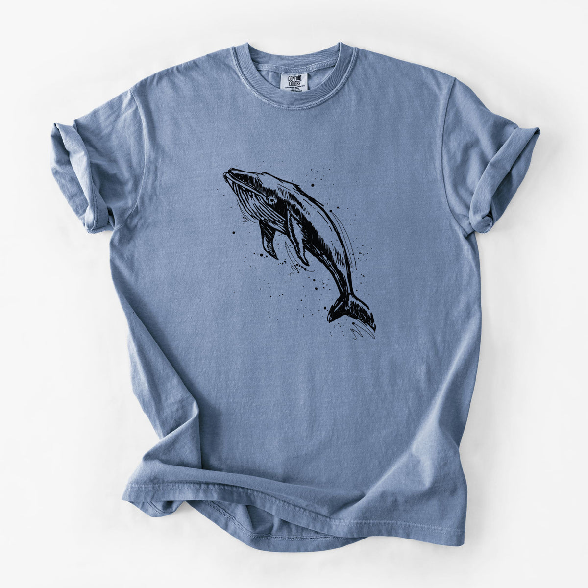 Blue jean Comfort Colors t-shirt laid flat, printed with a black ink illustration of a humpback whale diving, with ink splatters.
