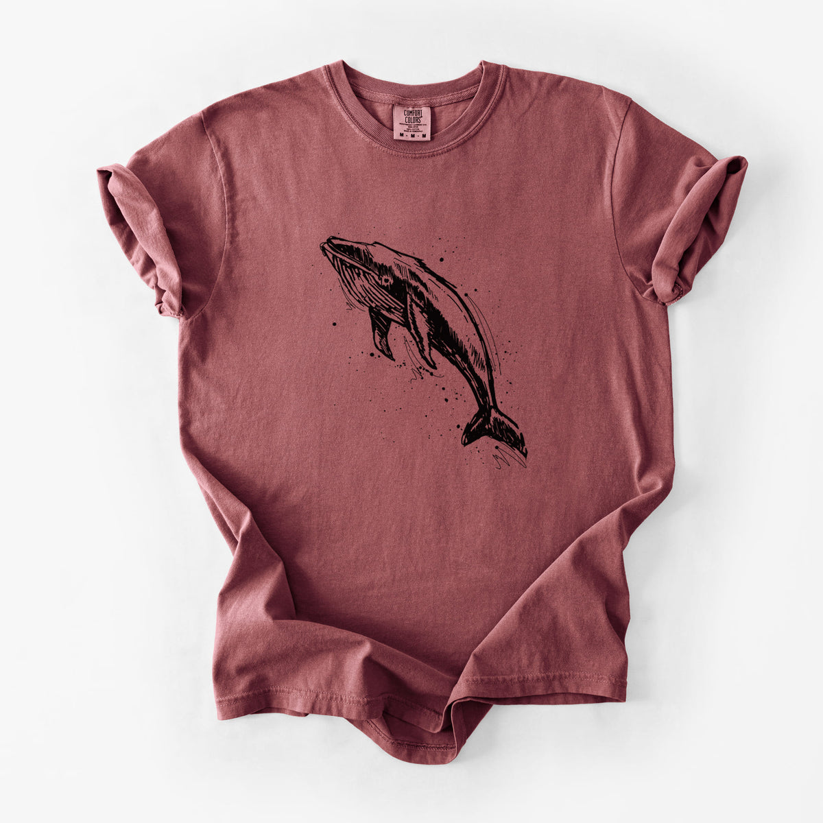 Brick red Comfort Colors t-shirt laid flat, printed with a black ink illustration of a humpback whale diving, with ink splatters.