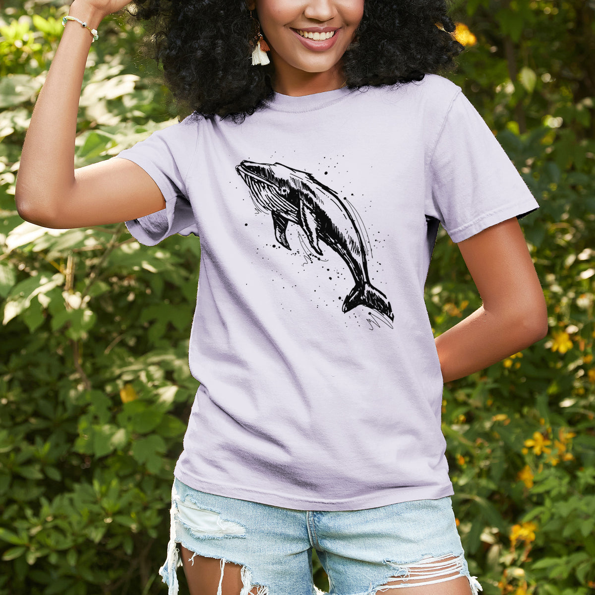Woman wearing a Comfort Colors t-shirt printed with a black ink illustration of a humpback whale diving, with ink splatters, outdoors.