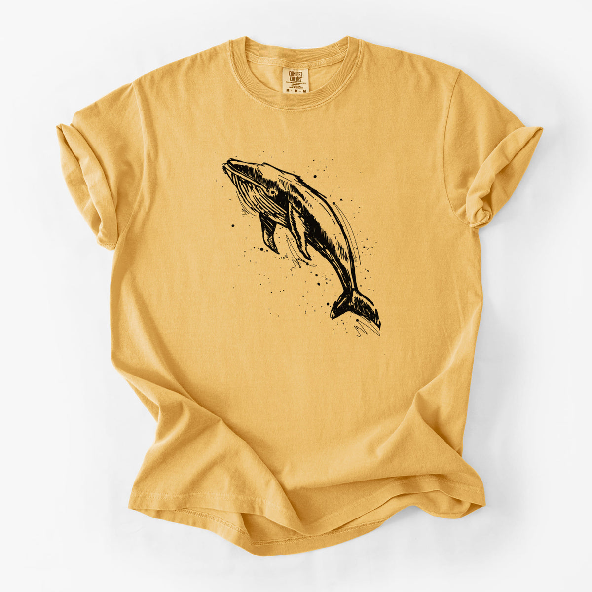 Golden wheat yellow Comfort Colors t-shirt laid flat, printed with a black ink illustration of a humpback whale diving, with ink splatters.
