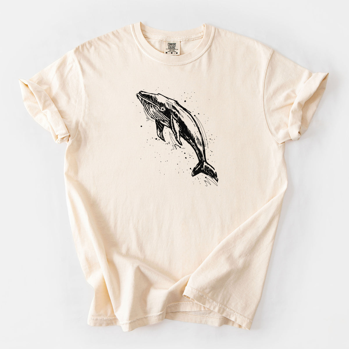 Ivory Comfort Colors t-shirt laid flat, printed with a black ink illustration of a humpback whale diving, with ink splatters.