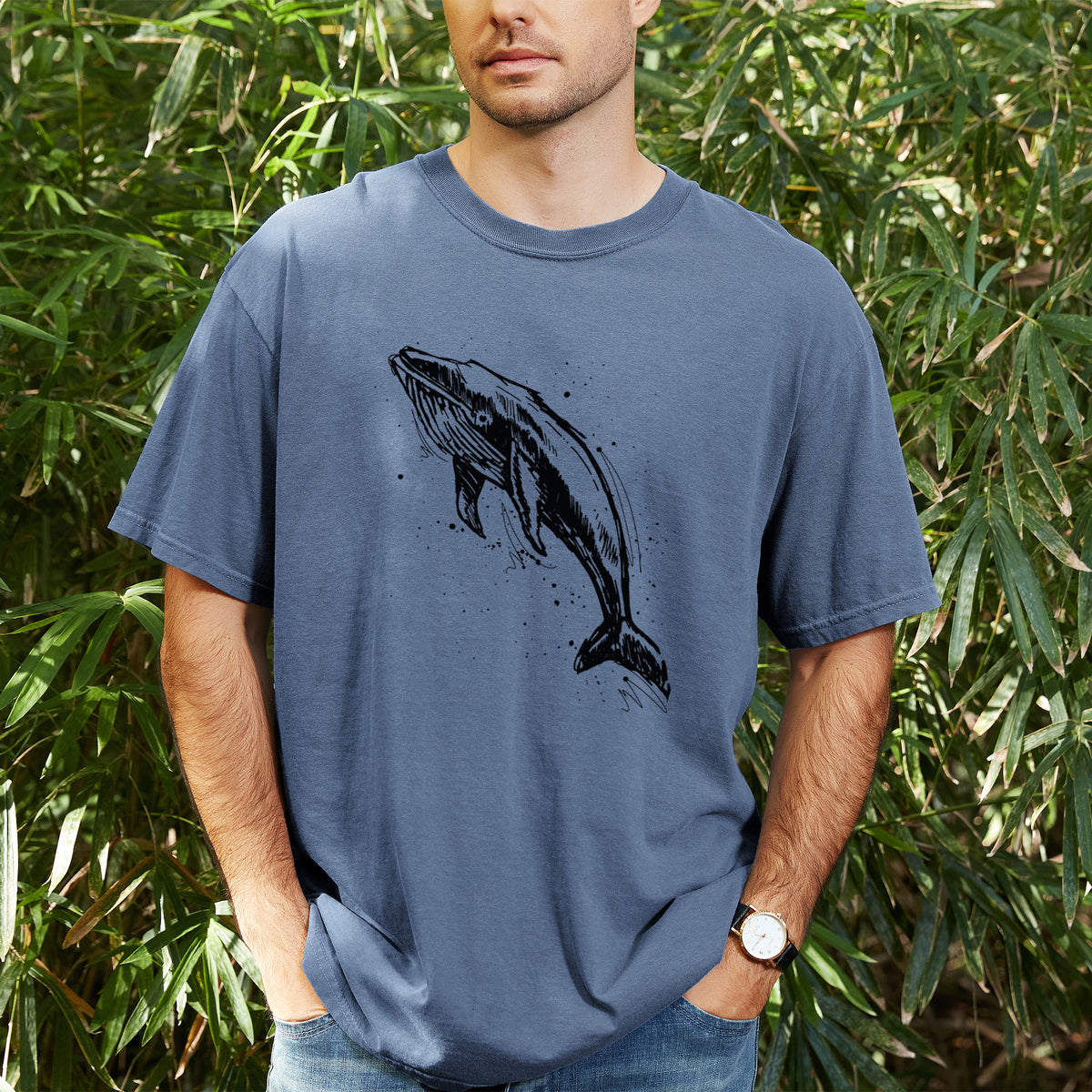 Man wearing a Comfort Colors t-shirt printed with a black ink illustration of a humpback whale diving, with ink splatters, outdoors.