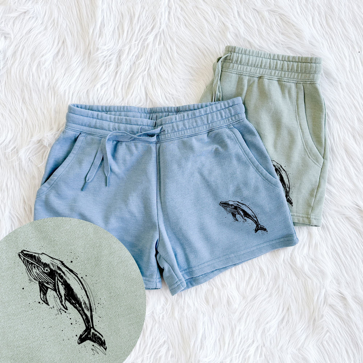 Two pairs of Cali Wave shorts in different colors printed with a black ink illustration of a humpback whale diving, with ink splatters, with a close-up of the print.