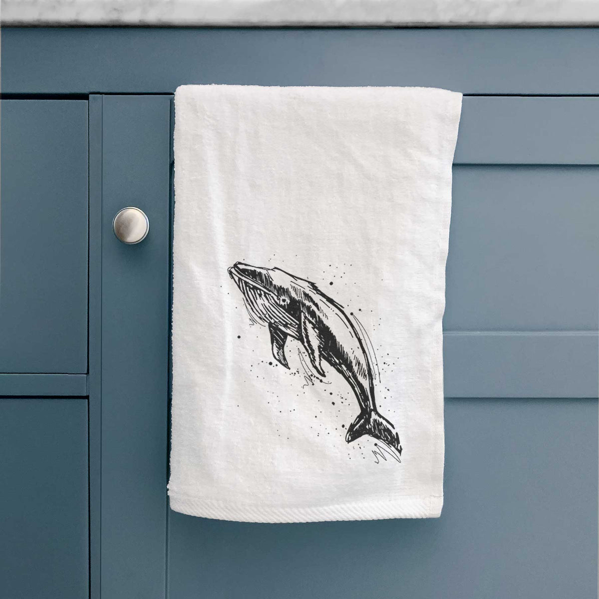 White hand towel printed with a black ink illustration of a humpback whale diving, with ink splatters, hanging on a cabinet door.