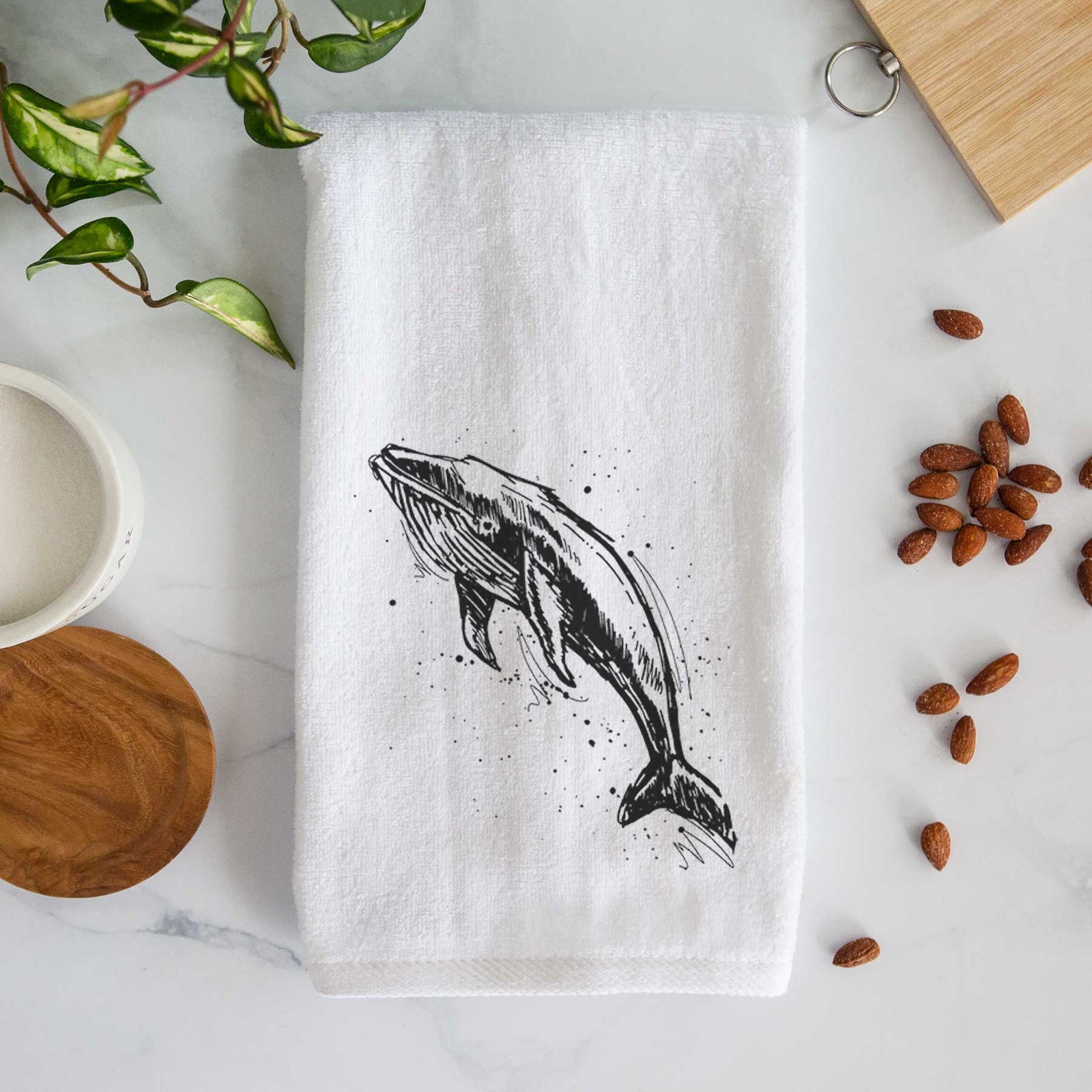 White hand towel printed with a black ink illustration of a humpback whale diving, with ink splatters.