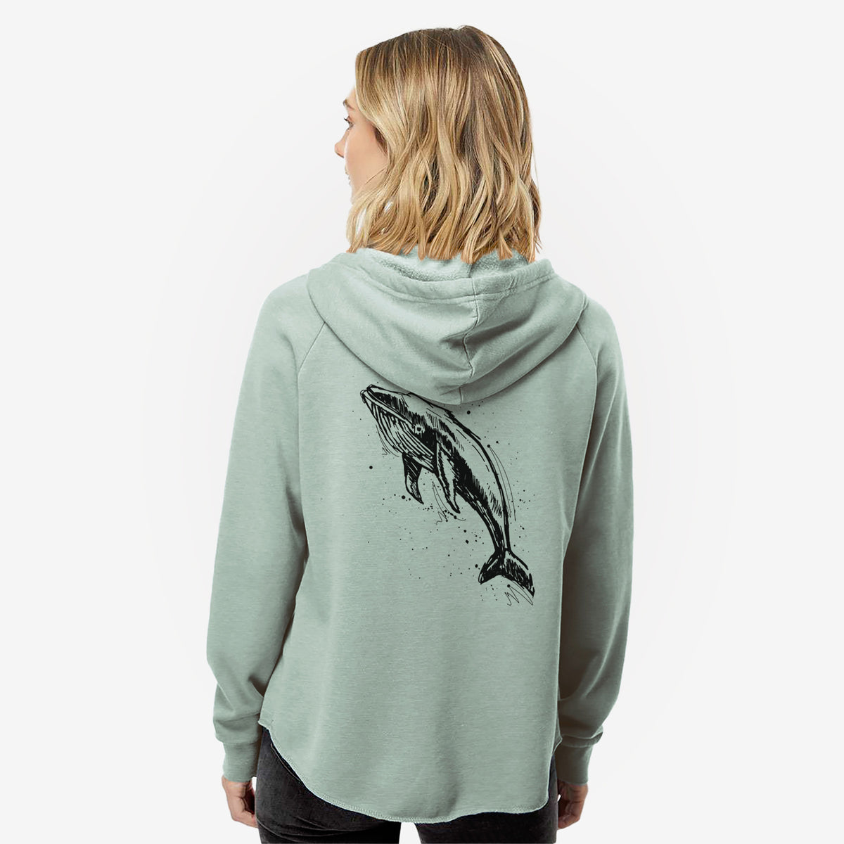 Woman wearing a sage green women's Cali Wave zip-up sweatshirt printed with a black ink illustration of a humpback whale diving, with ink splatters, seen from behind.