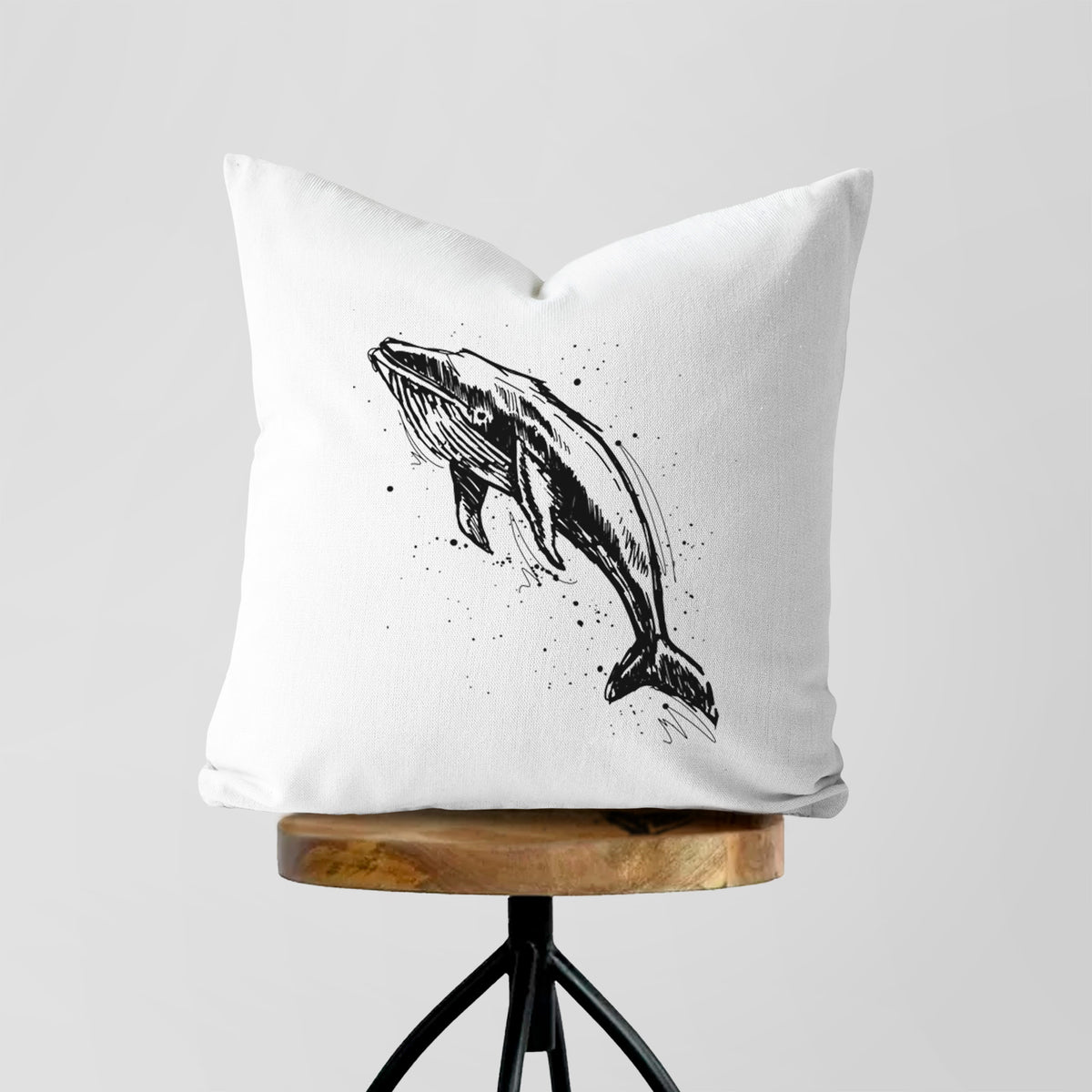 White pillow cover printed with a black ink illustration of a humpback whale diving, with ink splatters, on a wooden stool.