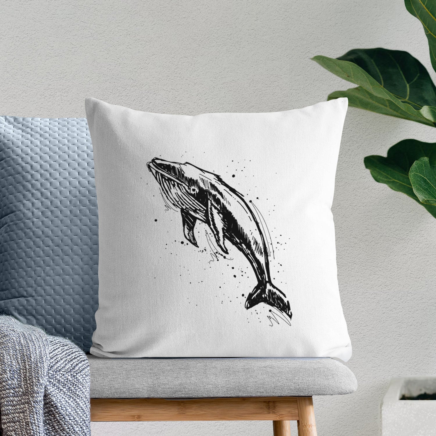 White pillow cover printed with a black ink illustration of a humpback whale diving, with ink splatters, on a bench beside plants.