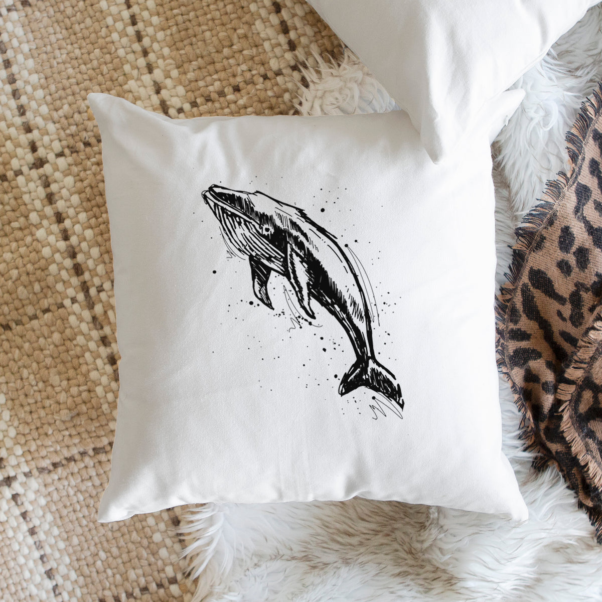 White pillow cover printed with a black ink illustration of a humpback whale diving, with ink splatters, on a woven chair.