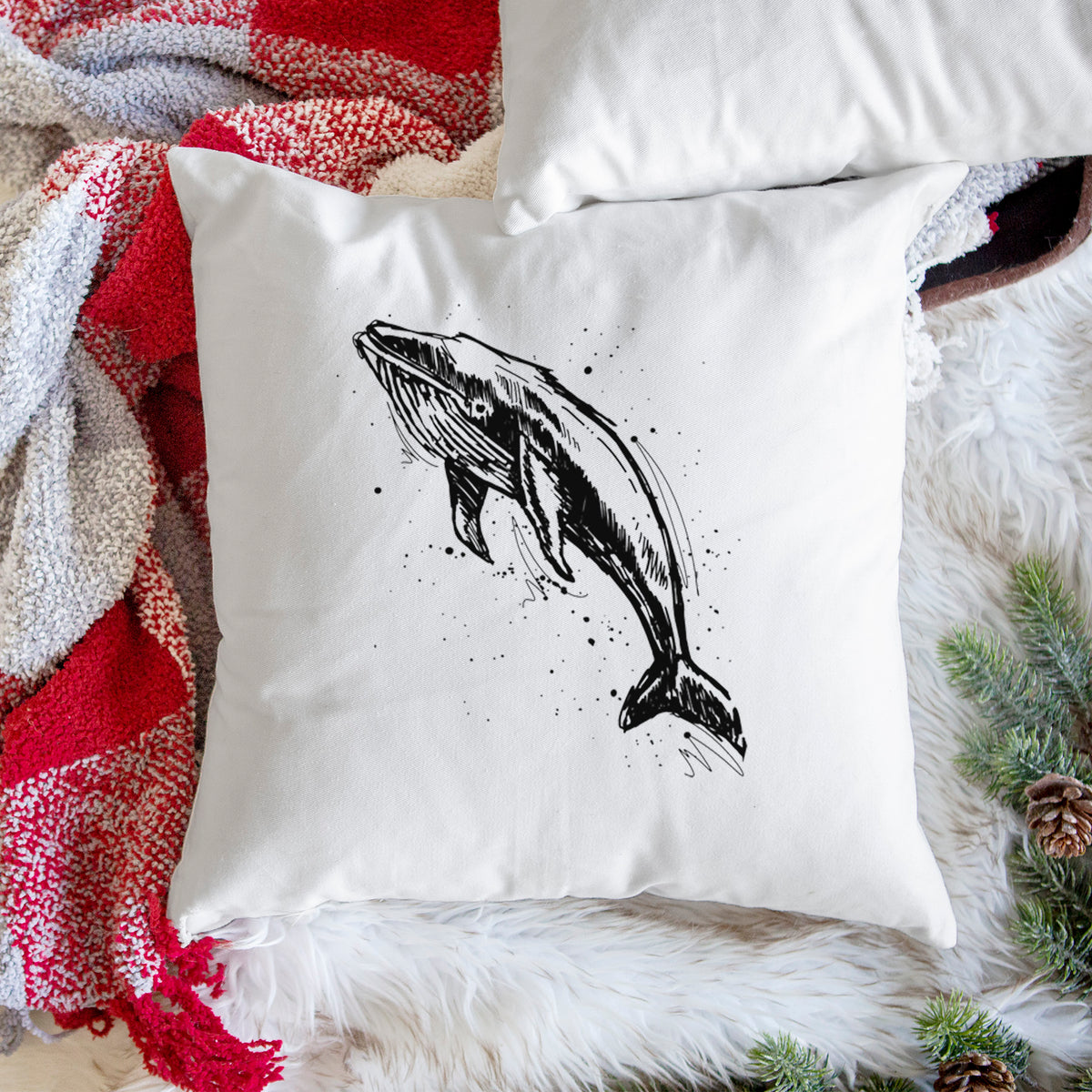 White pillow cover printed with a black ink illustration of a humpback whale diving, with ink splatters, on a woven chair.