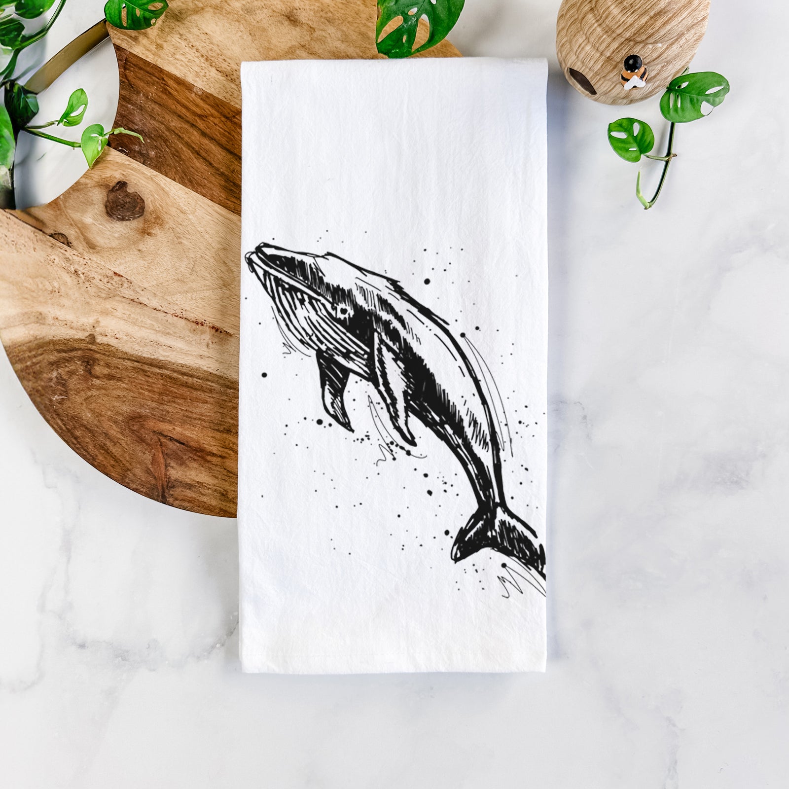 White cotton tea towel printed with a black ink illustration of a humpback whale diving, with ink splatters, on a marble counter beside a round wooden cutting board and greenery.