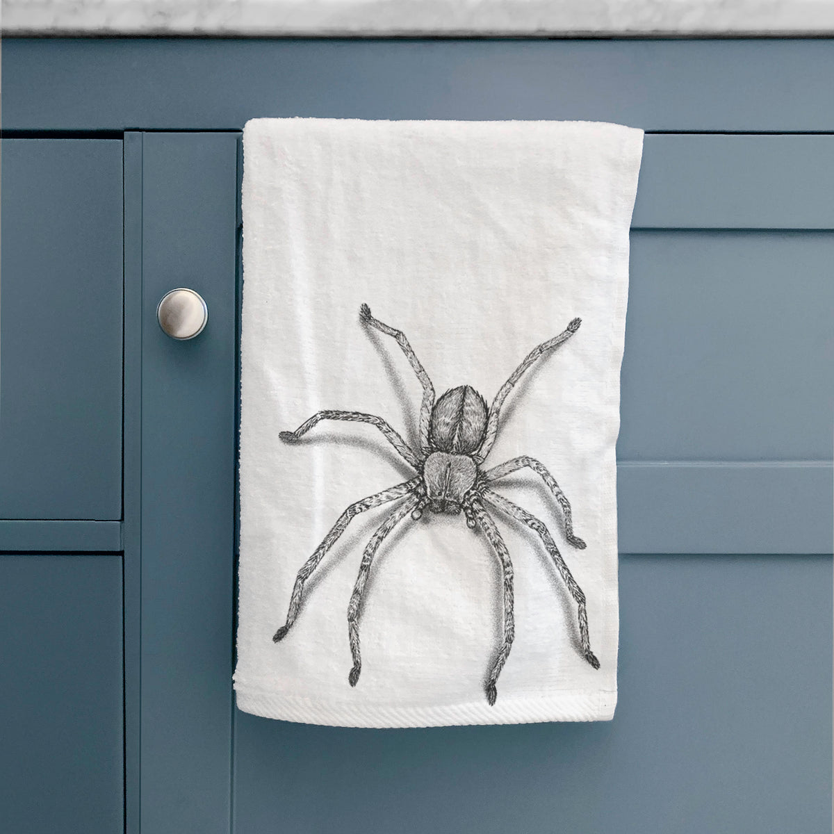 White hand towel printed with a detailed black ink illustration of a huntsman spider with long legs spread, hanging on a cabinet door.