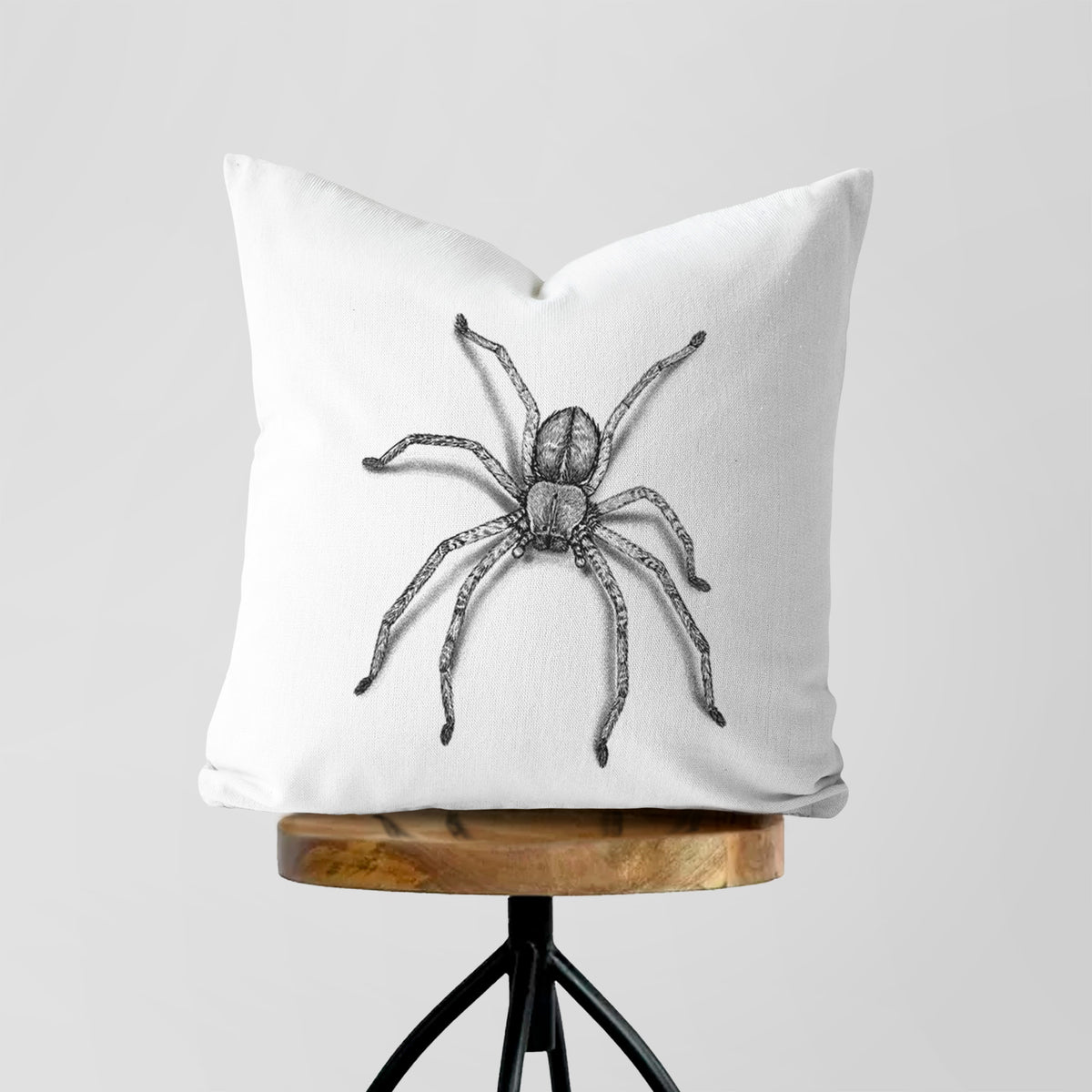 White pillow cover printed with a detailed black ink illustration of a huntsman spider with long legs spread, on a wooden stool.