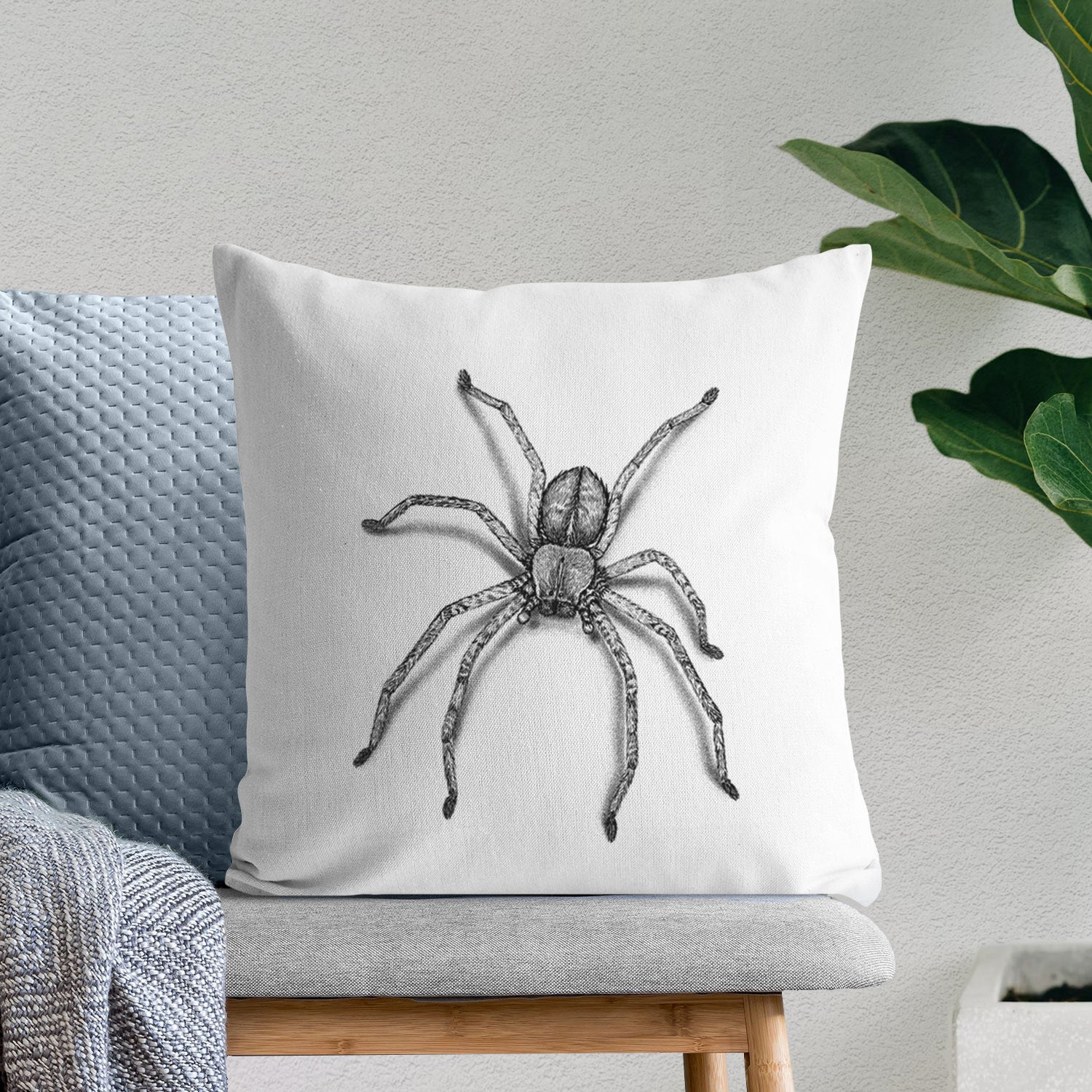 White pillow cover printed with a detailed black ink illustration of a huntsman spider with long legs spread, on a bench beside plants.