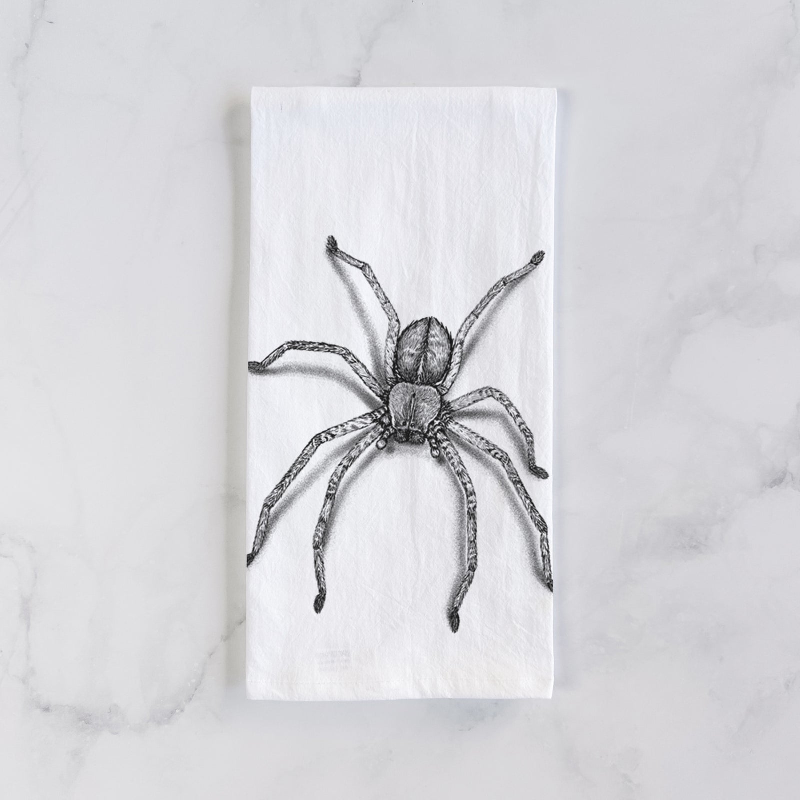 White cotton tea towel printed with a detailed black ink illustration of a huntsman spider with long legs spread, on a marble counter beside a round wooden cutting board and greenery.