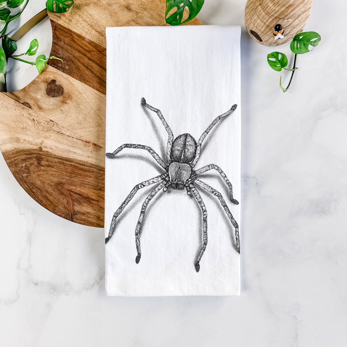 White cotton tea towel printed with a detailed black ink illustration of a huntsman spider with long legs spread, on a marble counter beside a round wooden cutting board and greenery.