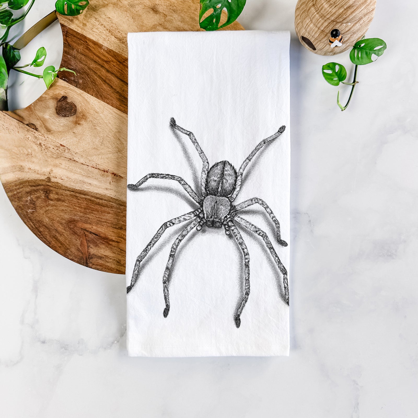 White cotton tea towel printed with a detailed black ink illustration of a huntsman spider with long legs spread, on a marble counter beside a round wooden cutting board and greenery.