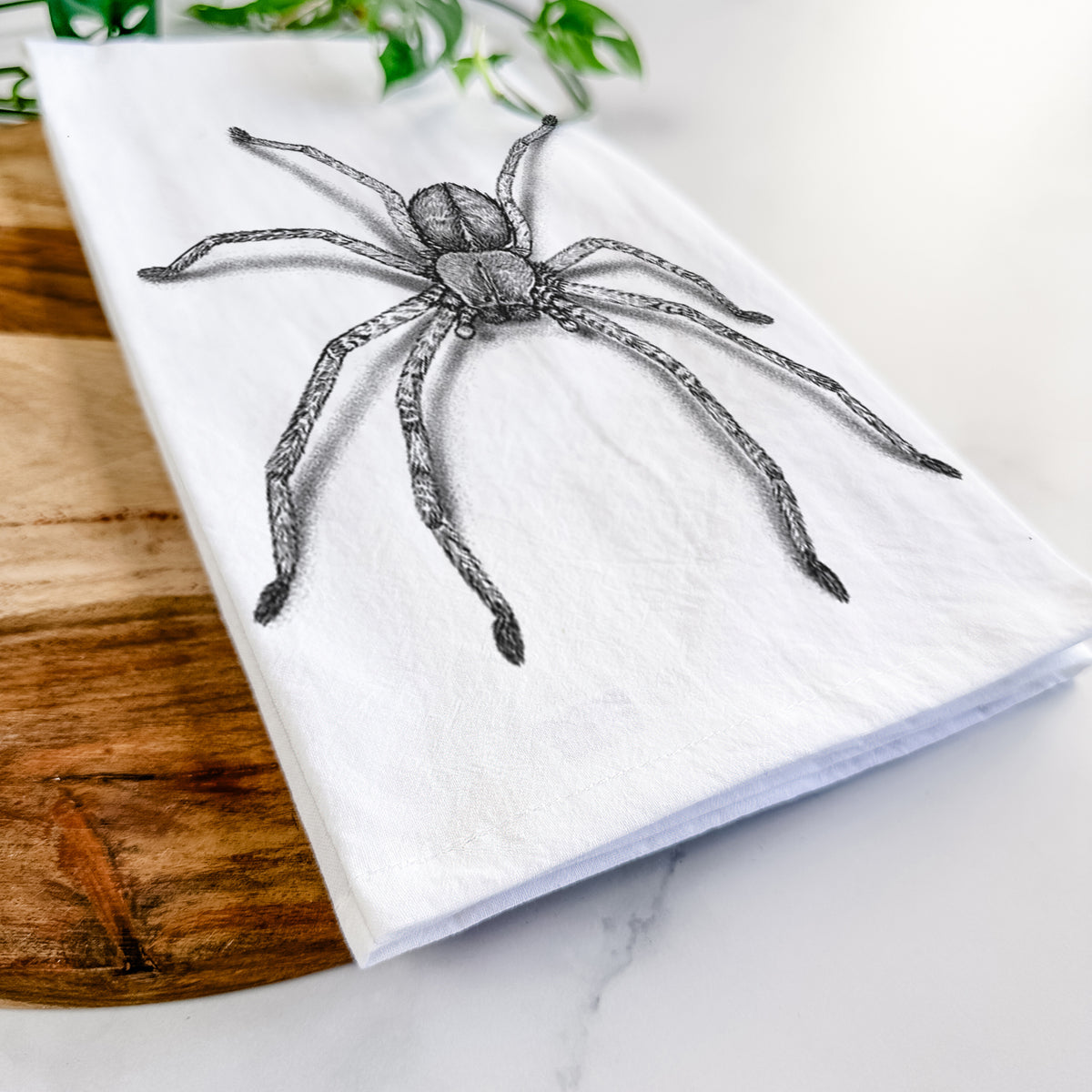 Close-up of a folded white tea towel on a wooden board, showing a detailed black ink illustration of a huntsman spider with long legs spread.