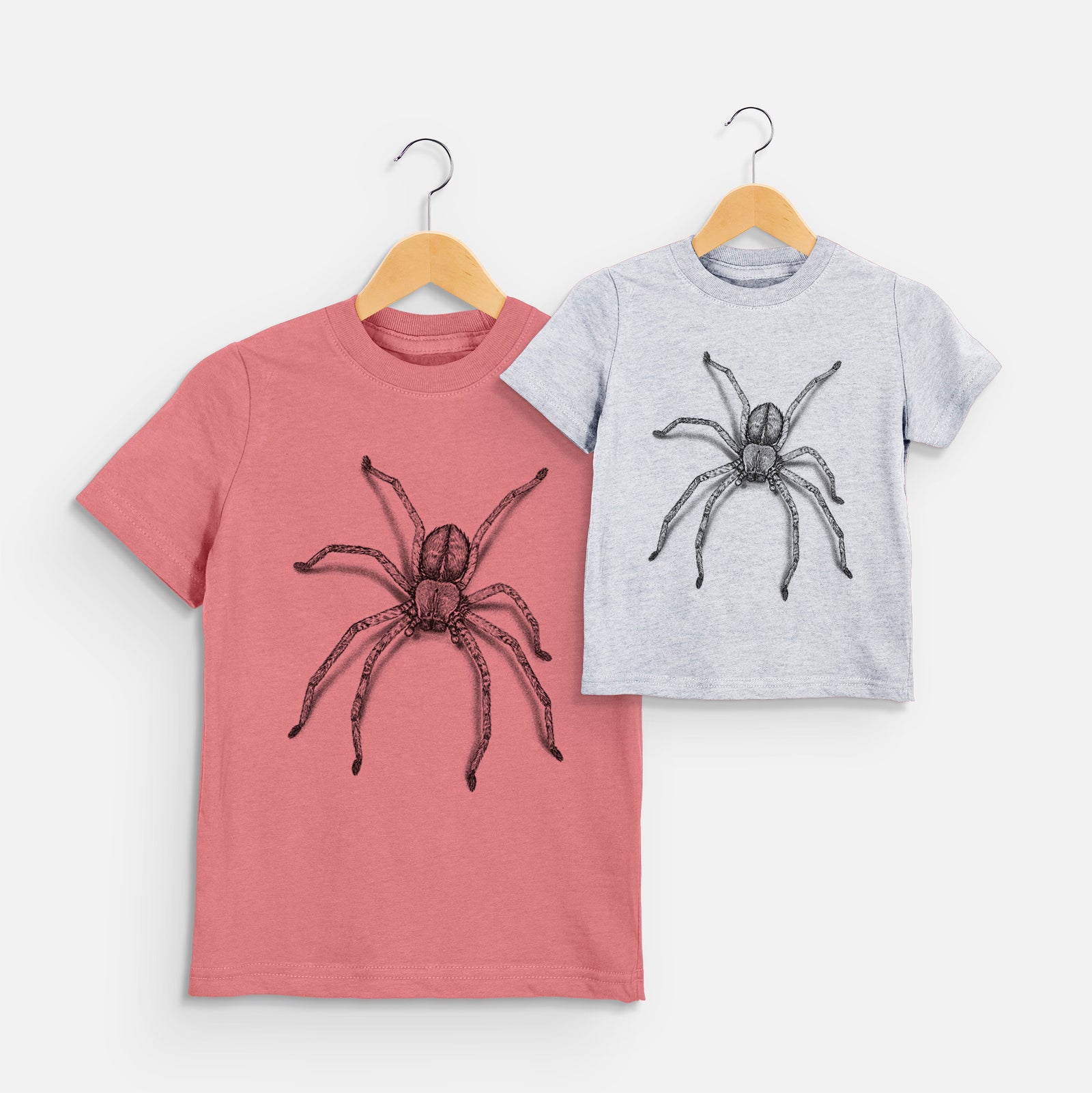 Two youth t-shirts in different colors on hangers, printed with a detailed black ink illustration of a huntsman spider with long legs spread.