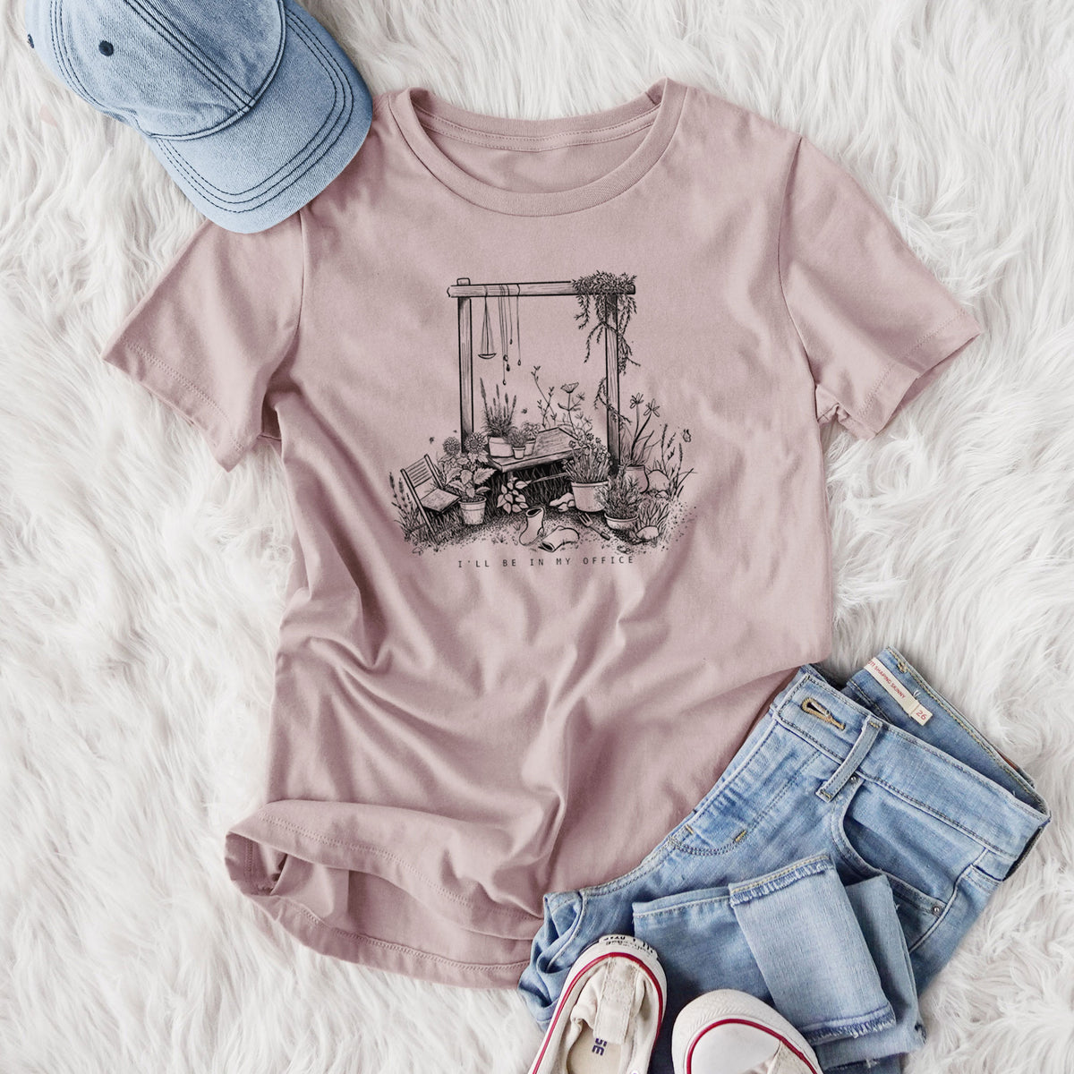 Women's relaxed fit cotton t-shirt printed with a black ink illustration of a garden gate arch surrounded by potted plants and flowers, styled flat on a white faux fur rug with jeans.