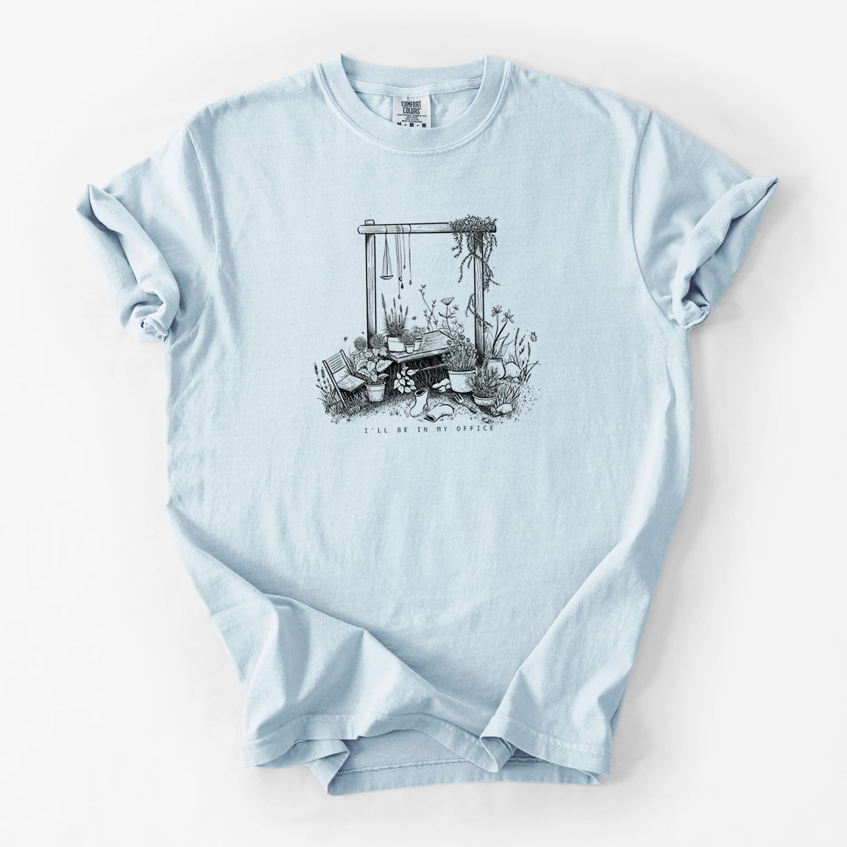 Chambray blue Comfort Colors t-shirt laid flat, printed with a black ink illustration of a garden gate arch surrounded by potted plants and flowers.
