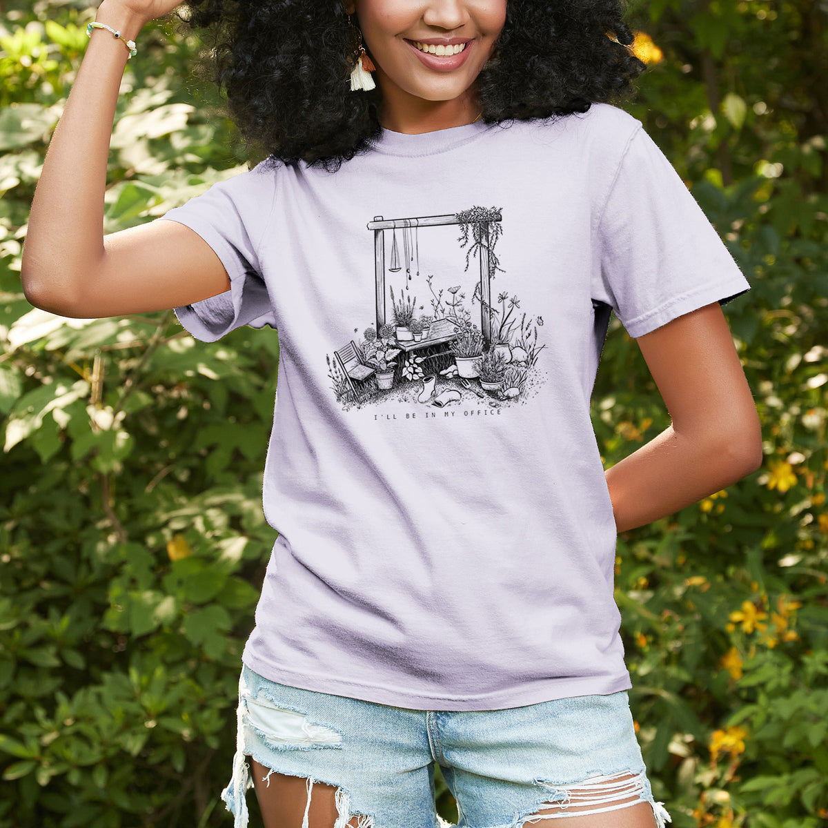 Woman wearing a Comfort Colors t-shirt printed with a black ink illustration of a garden gate arch surrounded by potted plants and flowers, outdoors.
