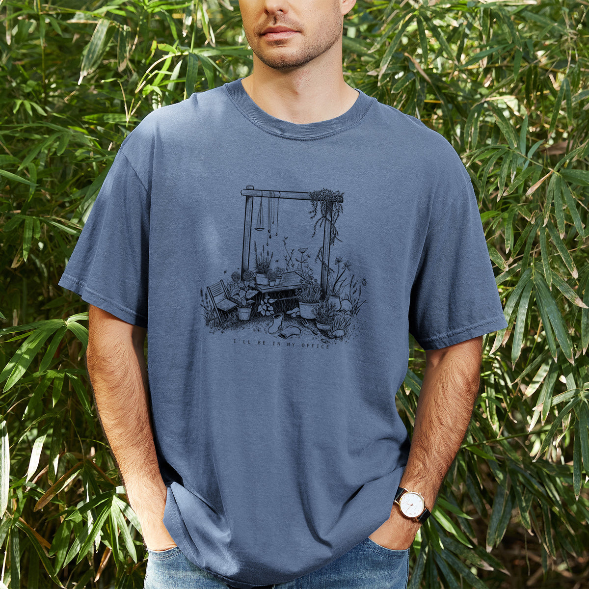 Man wearing a Comfort Colors t-shirt printed with a black ink illustration of a garden gate arch surrounded by potted plants and flowers, outdoors.
