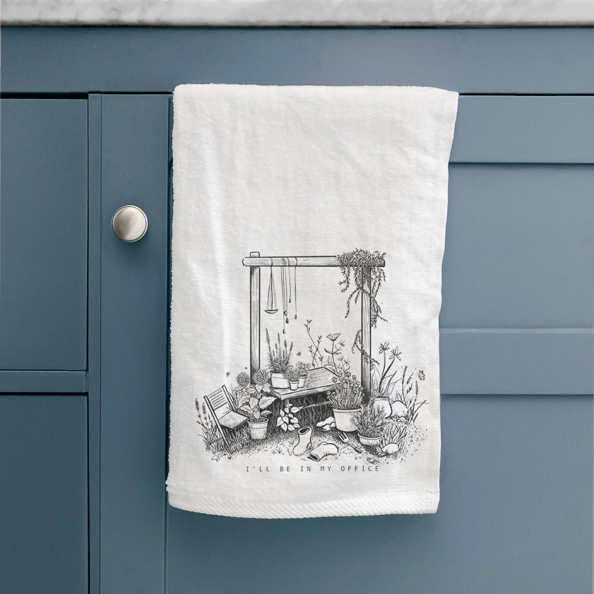 White hand towel printed with a black ink illustration of a garden gate arch surrounded by potted plants and flowers, hanging on a cabinet door.