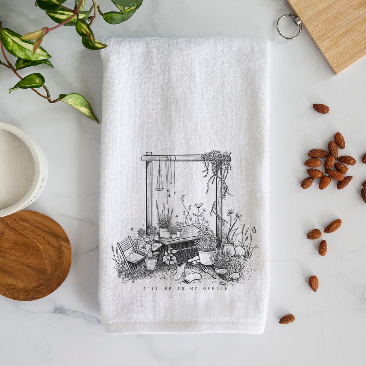White hand towel printed with a black ink illustration of a garden gate arch surrounded by potted plants and flowers.