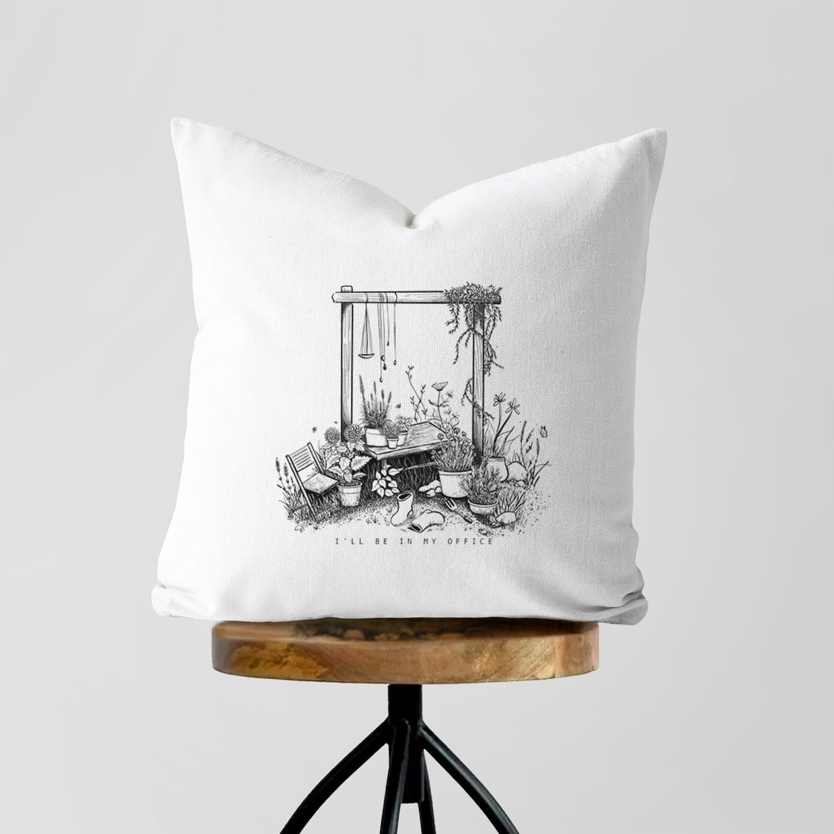 White pillow cover printed with a black ink illustration of a garden gate arch surrounded by potted plants and flowers, on a wooden stool.