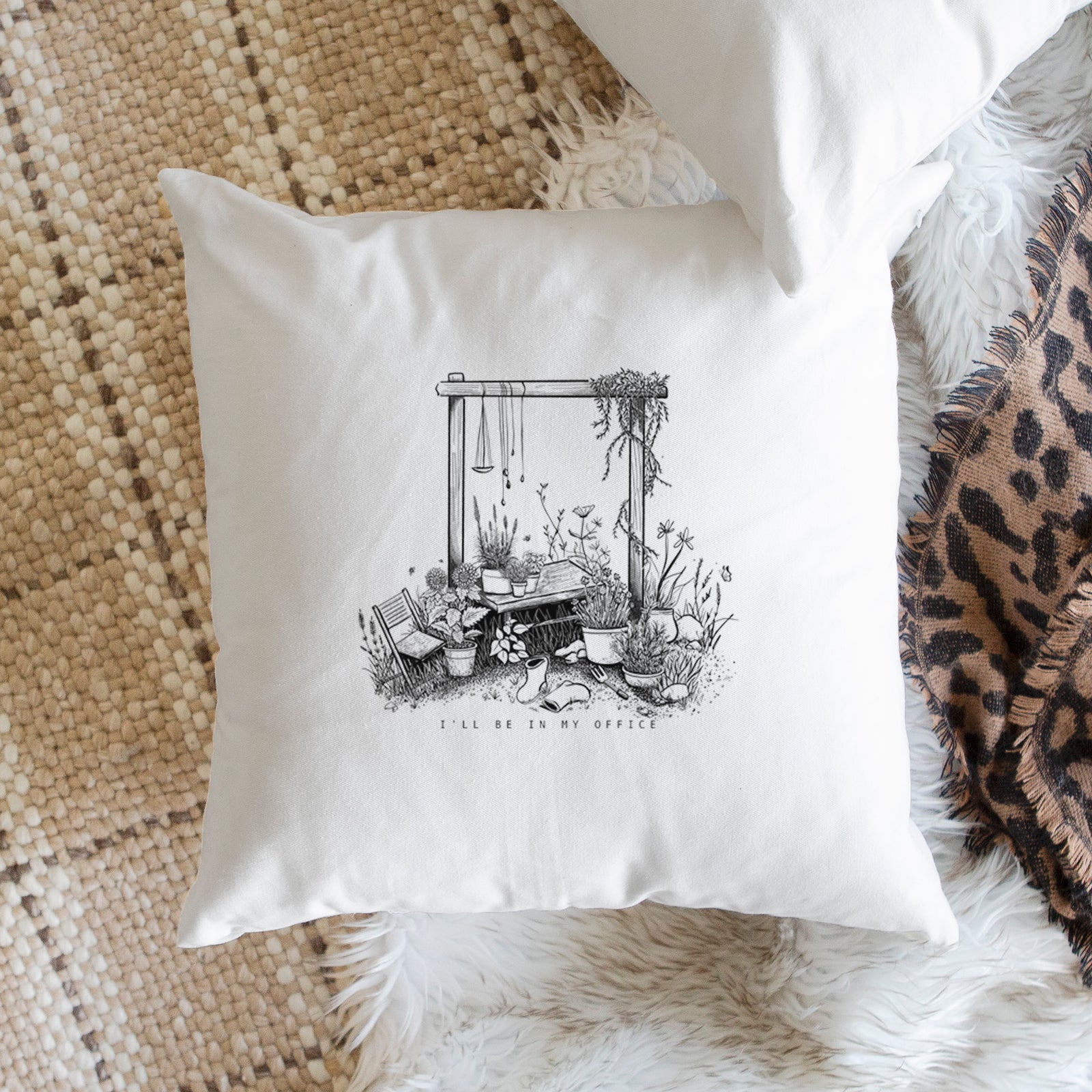 White pillow cover printed with a black ink illustration of a garden gate arch surrounded by potted plants and flowers, on a bench beside plants.