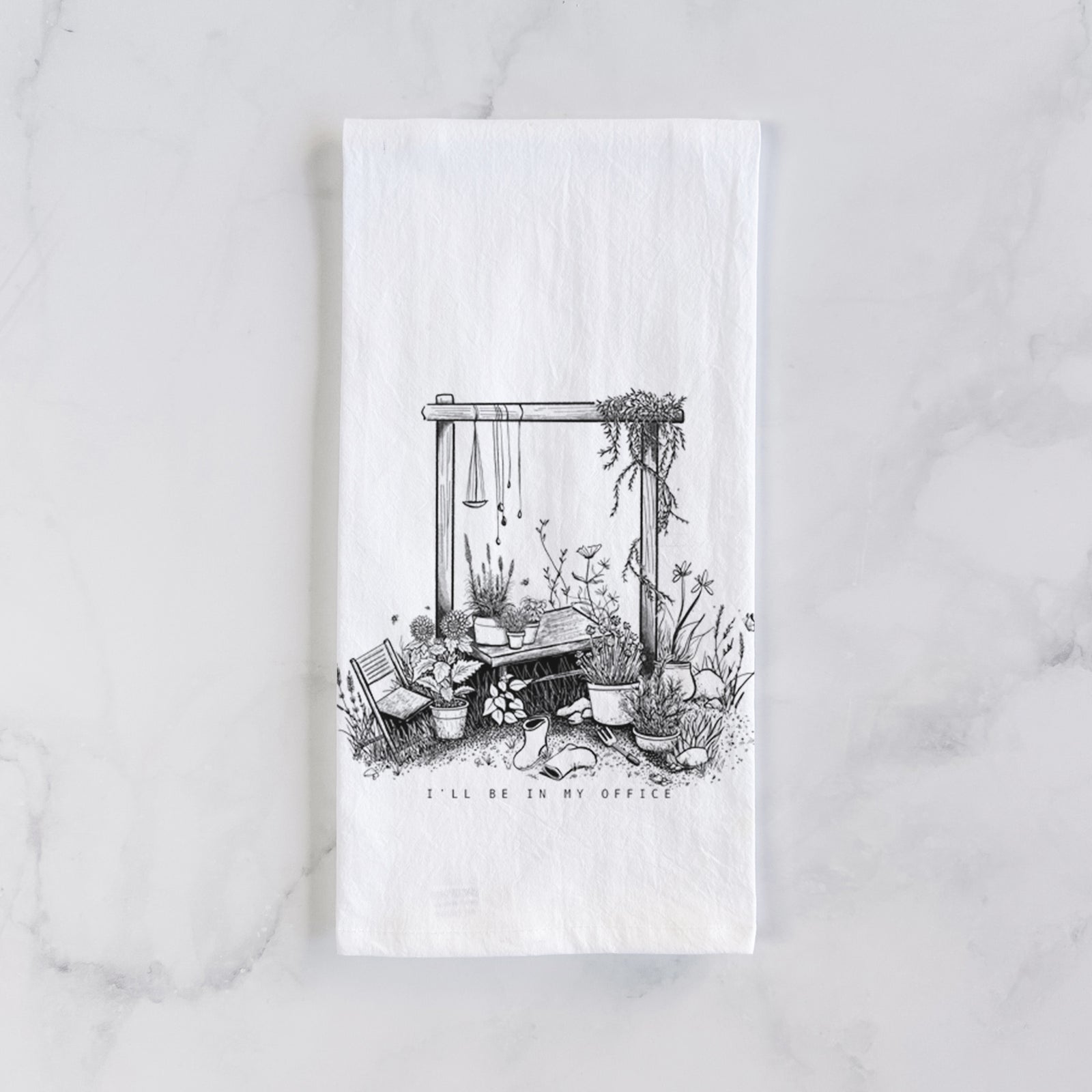 White cotton tea towel printed with a black ink illustration of a garden gate arch surrounded by potted plants and flowers, on a marble counter beside a round wooden cutting board and greenery.