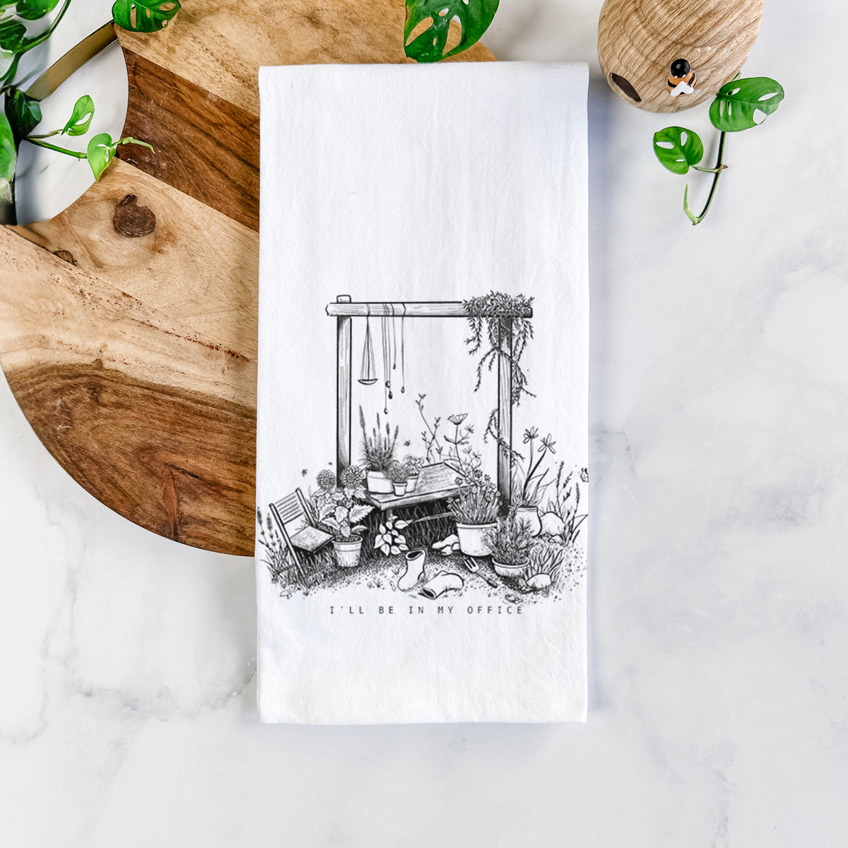White cotton tea towel printed with a black ink illustration of a garden gate arch surrounded by potted plants and flowers, on a marble counter beside a round wooden cutting board and greenery.