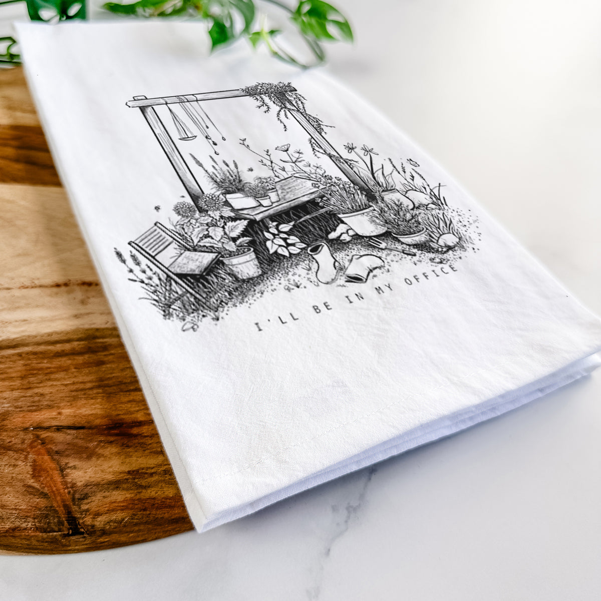 Close-up of a folded white tea towel on a wooden board, showing a black ink illustration of a garden gate arch surrounded by potted plants and flowers.
