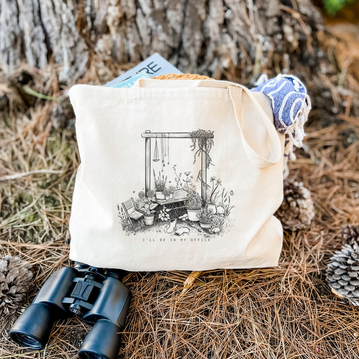 Canvas tote bag printed with a black ink illustration of a garden gate arch surrounded by potted plants and flowers, resting on dry grass beside a pair of binoculars.