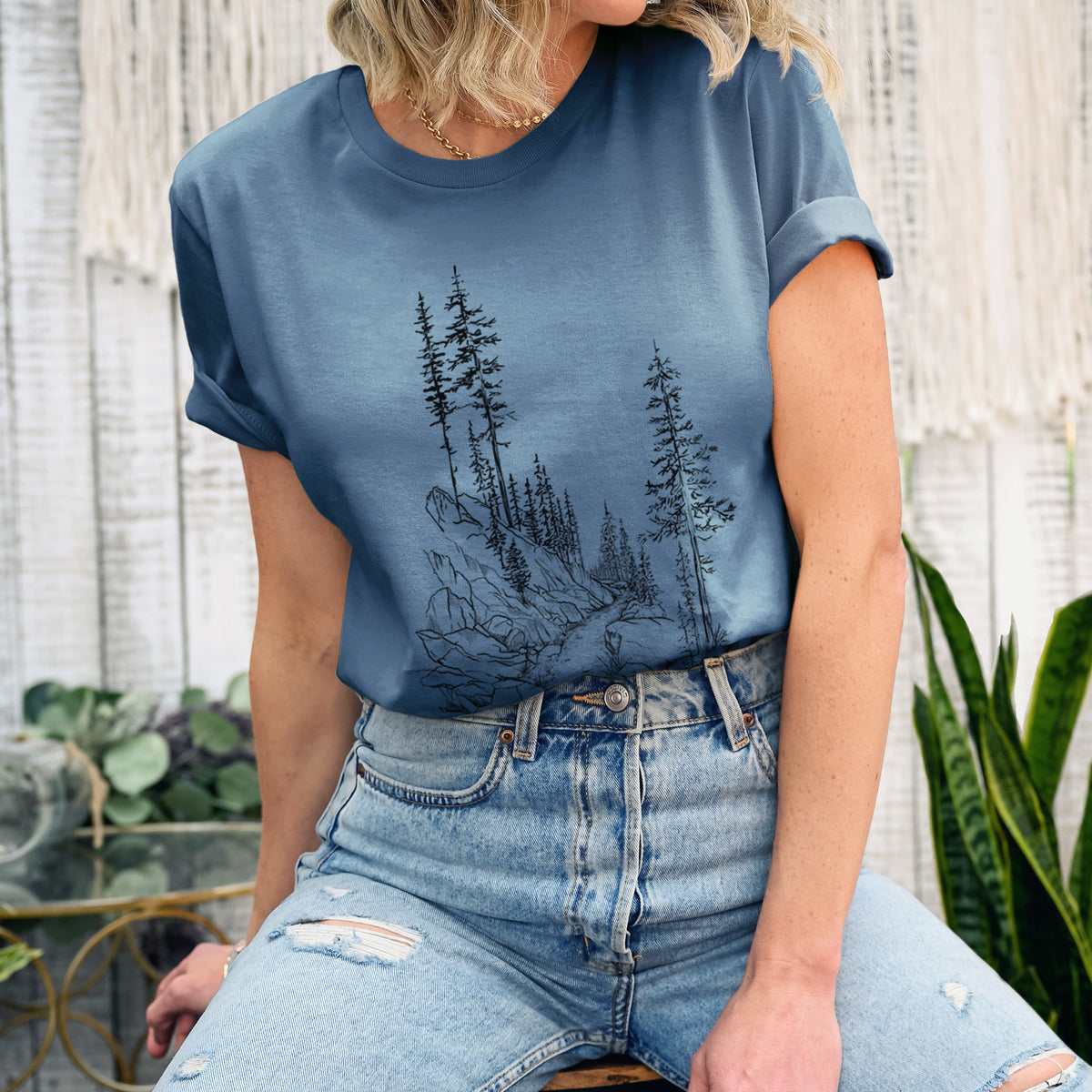 Woman wearing a steel blue lightweight cotton t-shirt printed with a black ink illustration of a trail winding past tall pine trees and rocks into the woods.