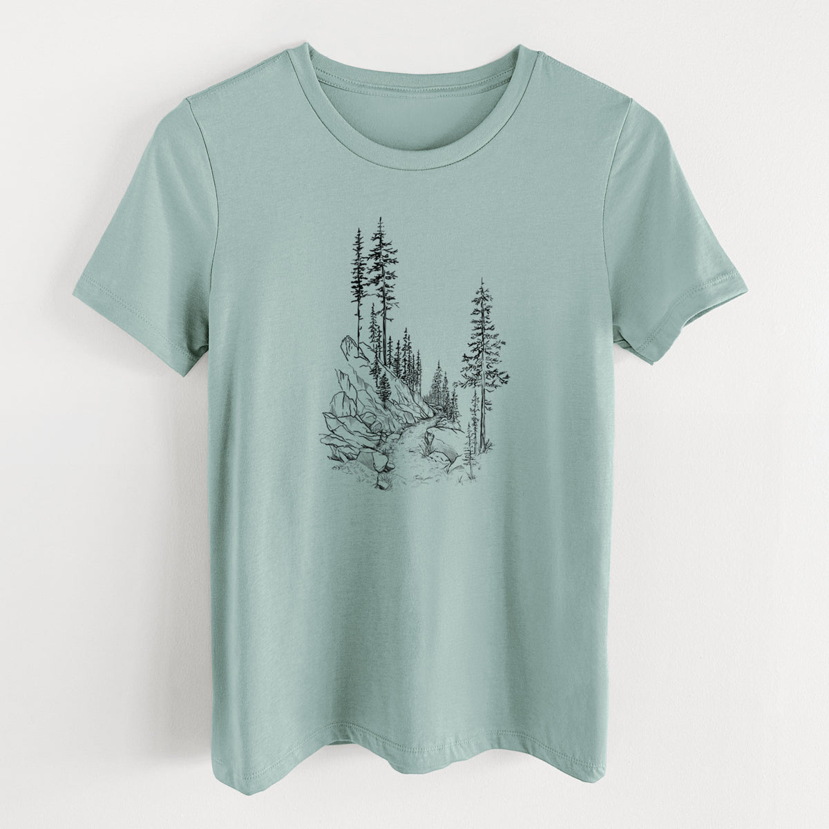 Dusty blue women's relaxed fit cotton t-shirt laid flat, printed with a black ink illustration of a trail winding past tall pine trees and rocks into the woods.