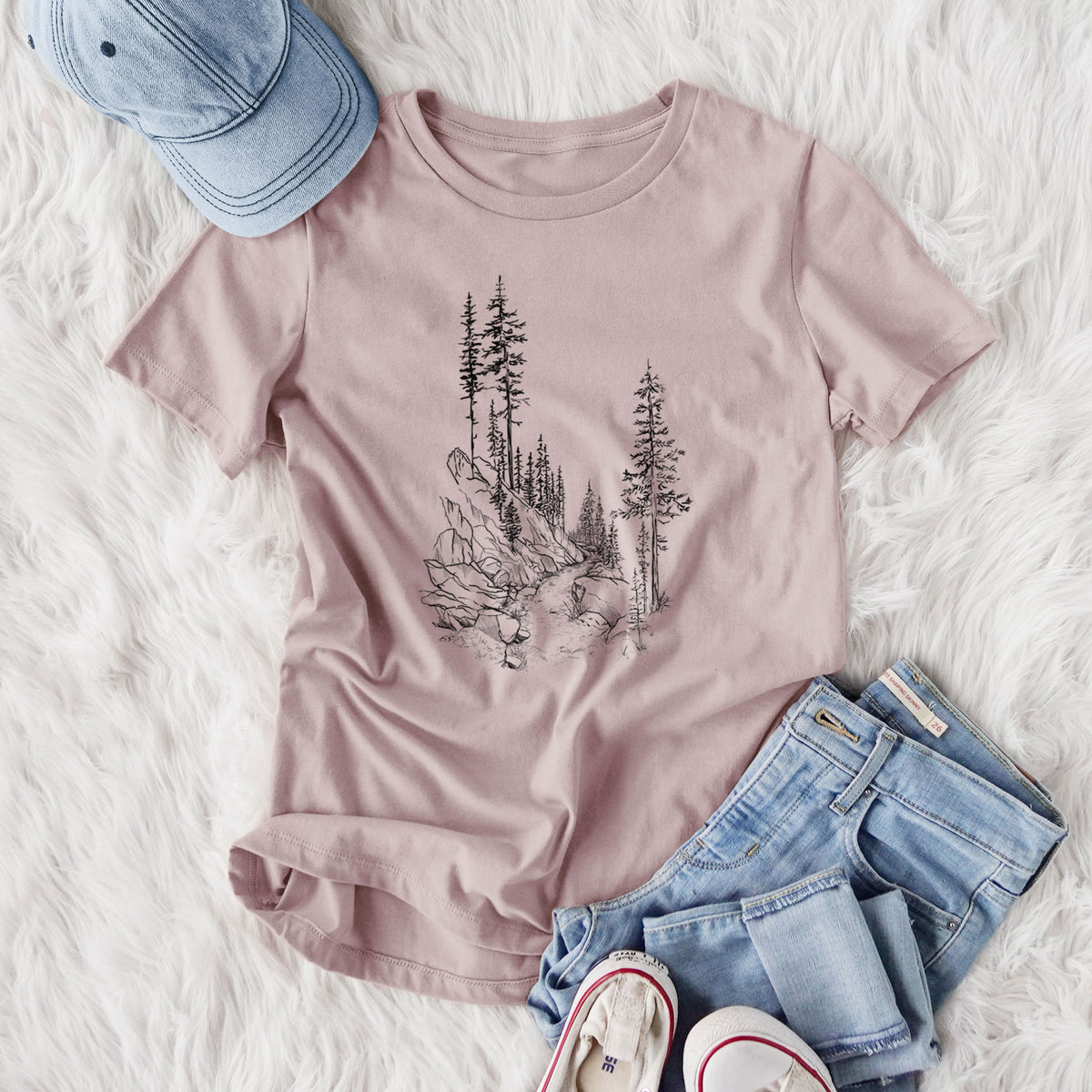 Women's relaxed fit cotton t-shirt printed with a black ink illustration of a trail winding past tall pine trees and rocks into the woods, styled flat on a white faux fur rug with jeans.
