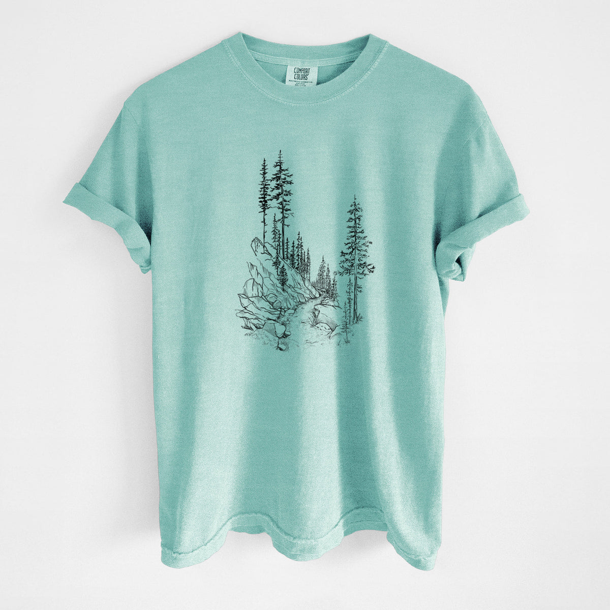 Chalky mint Comfort Colors t-shirt laid flat, printed with a black ink illustration of a trail winding past tall pine trees and rocks into the woods.
