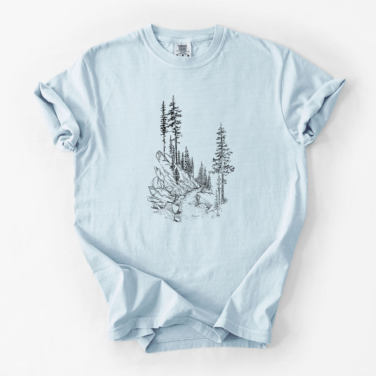 Chambray blue Comfort Colors t-shirt laid flat, printed with a black ink illustration of a trail winding past tall pine trees and rocks into the woods.