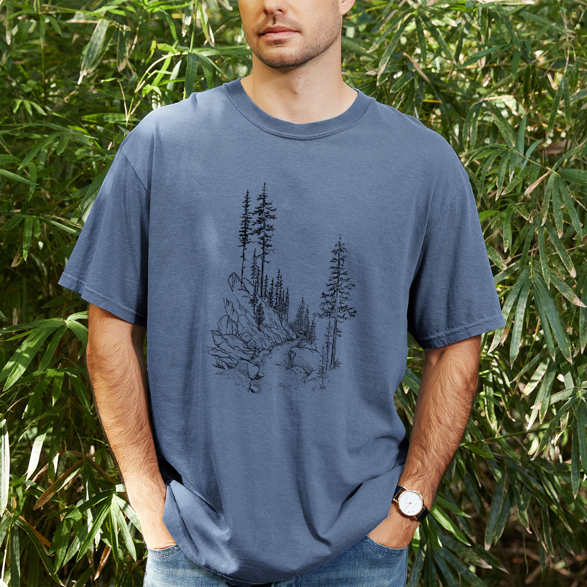 Man wearing a Comfort Colors t-shirt printed with a black ink illustration of a trail winding past tall pine trees and rocks into the woods, outdoors.