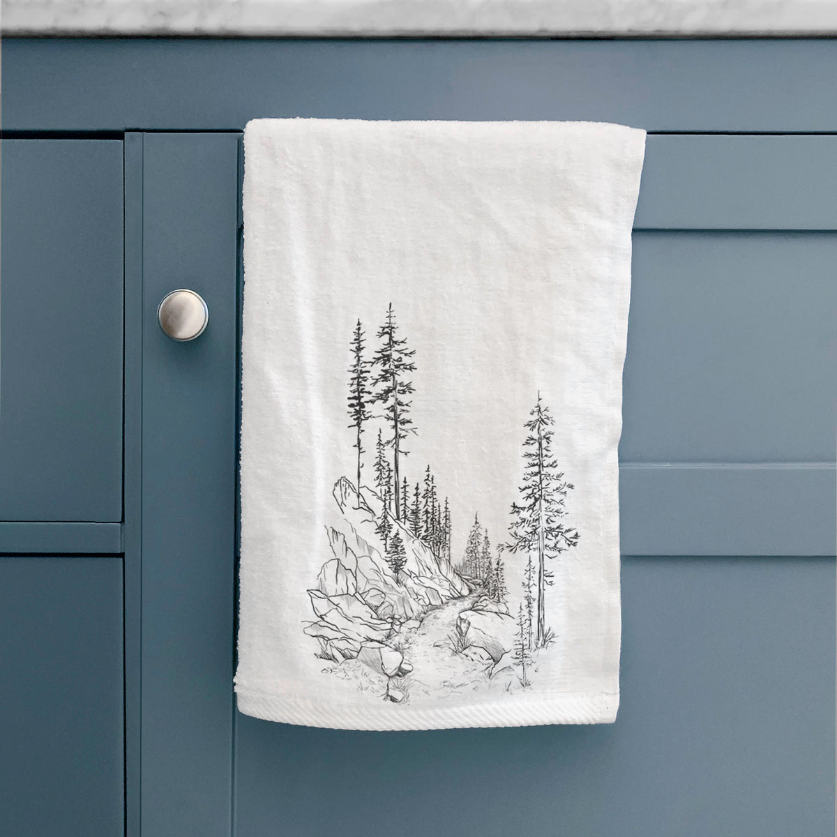 White hand towel printed with a black ink illustration of a trail winding past tall pine trees and rocks into the woods, hanging on a cabinet door.