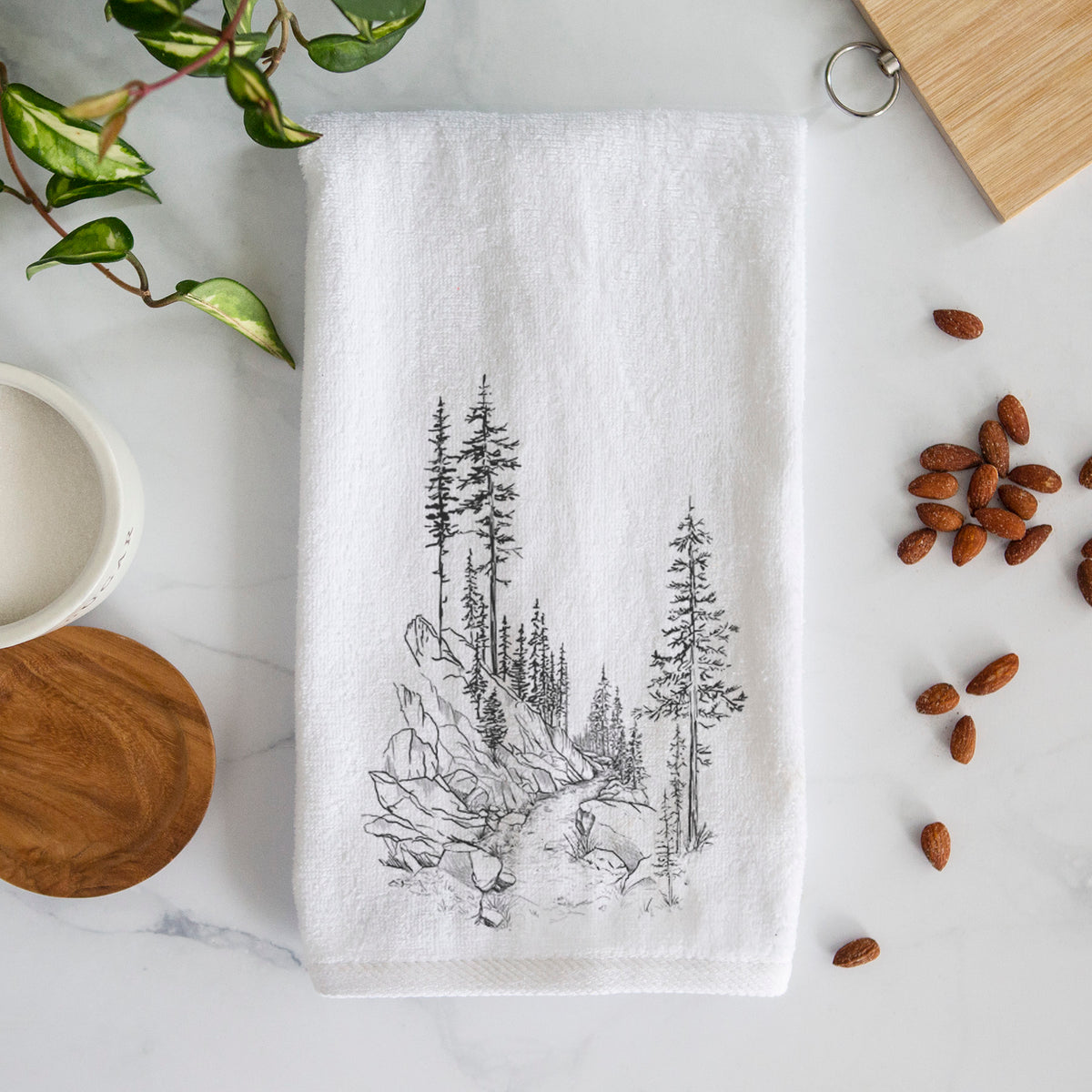 White hand towel printed with a black ink illustration of a trail winding past tall pine trees and rocks into the woods.
